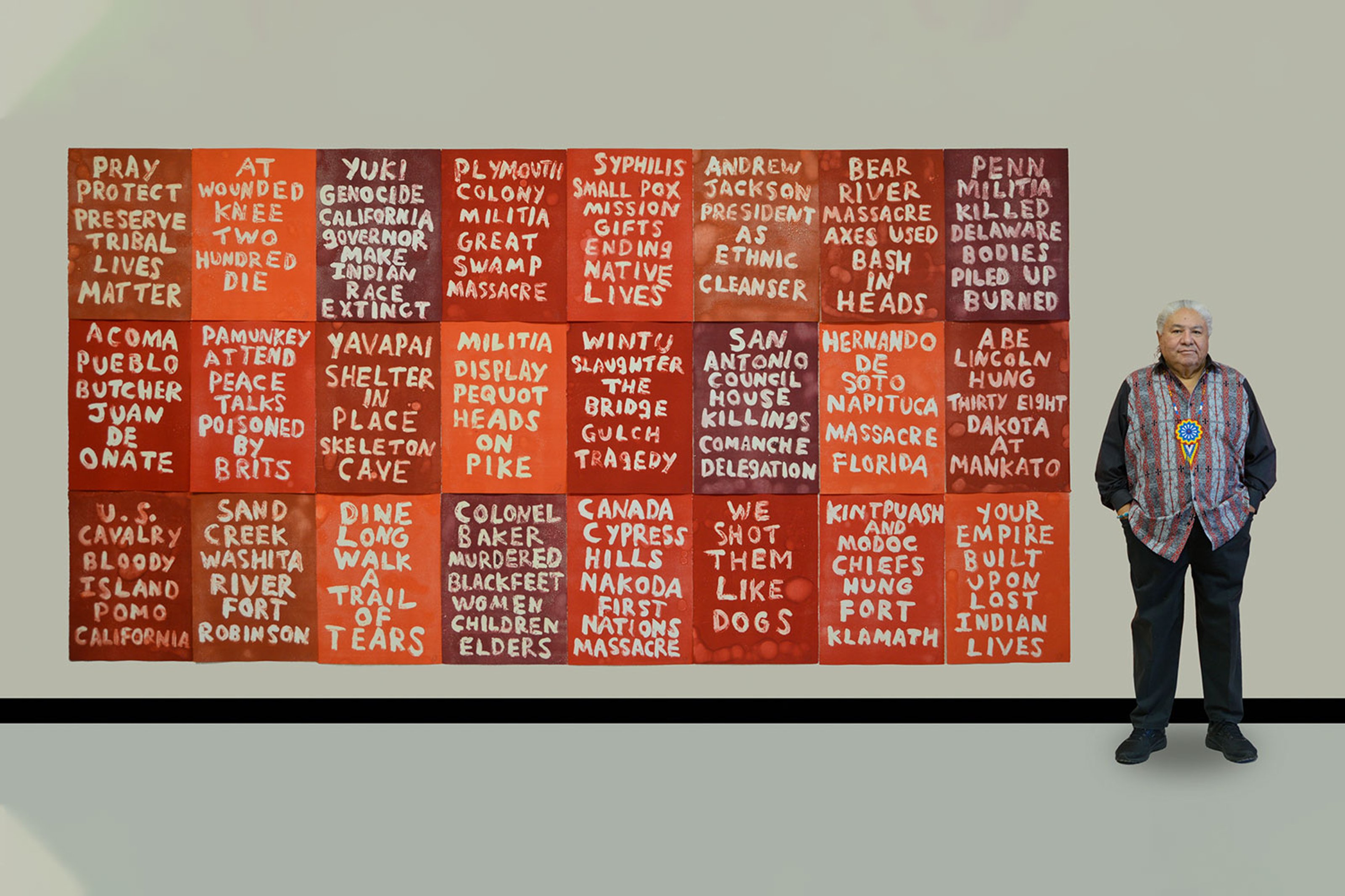 Artist Edgar Heap of Birds standing beside his artwork, which consists of multiple red and orange tiles with text highlighting historical injustices against Indigenous peoples.