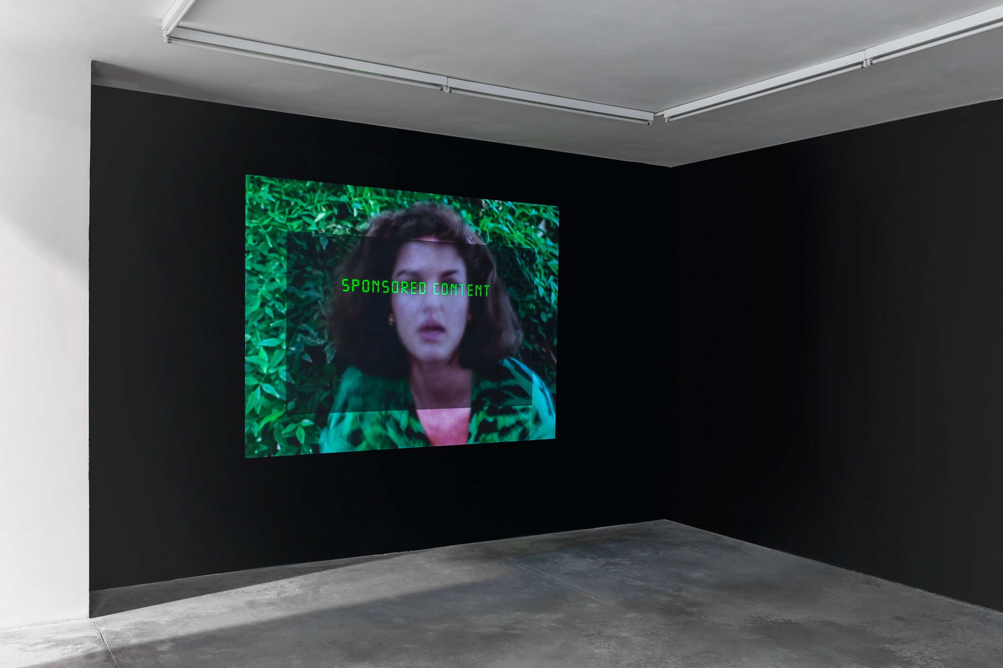 Large wall-mounted screen in an art gallery with black walls and a concrete floor, displaying an image of a black-haired woman in a green outfit with green leaves in the background. The words 'sponsored content' are visible on the screen.