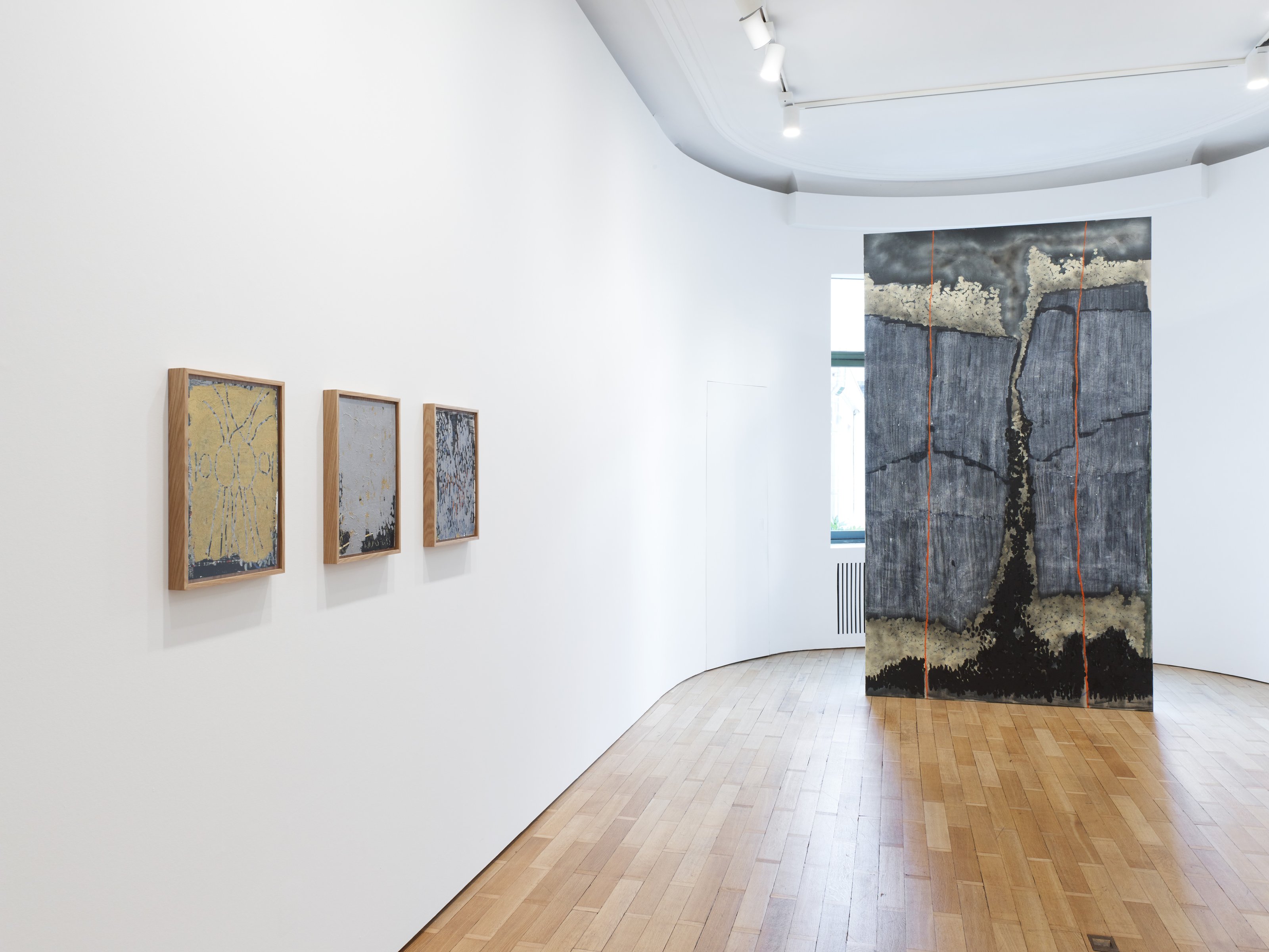 Three earth-toned patterned paintings in wooden frames hang on a white-walled, brightly lit art gallery wall, alongside a rectangular fabric with abstract earth-toned patterns hanging from the ceiling.