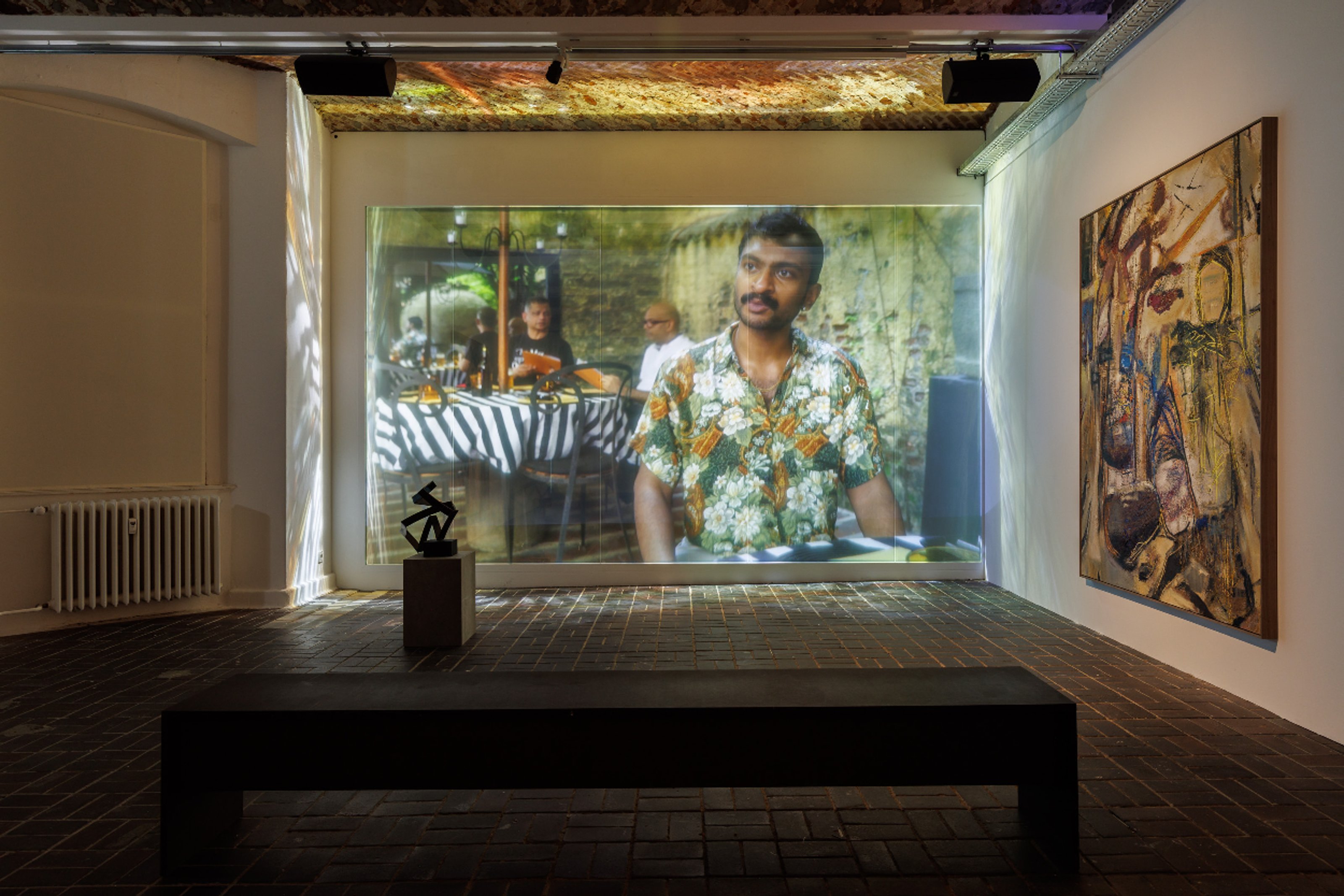 Video of a mustachioed man in a flowered shirt shown on a giant screen in an art gallery.