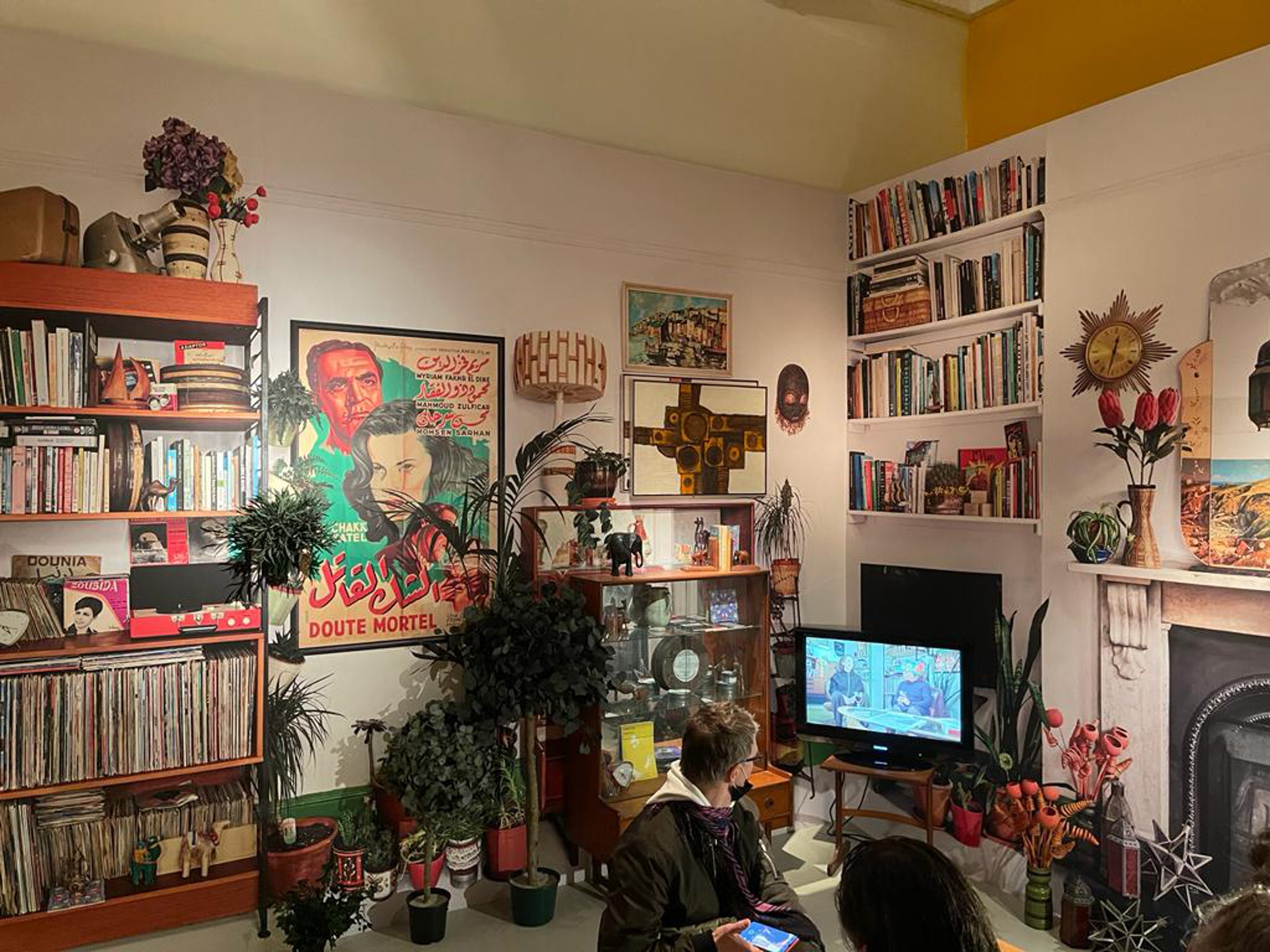 A person sitting on the floor looking at the TV over their shoulder in a room filled with books, paintings, and ornaments in yellow tones.