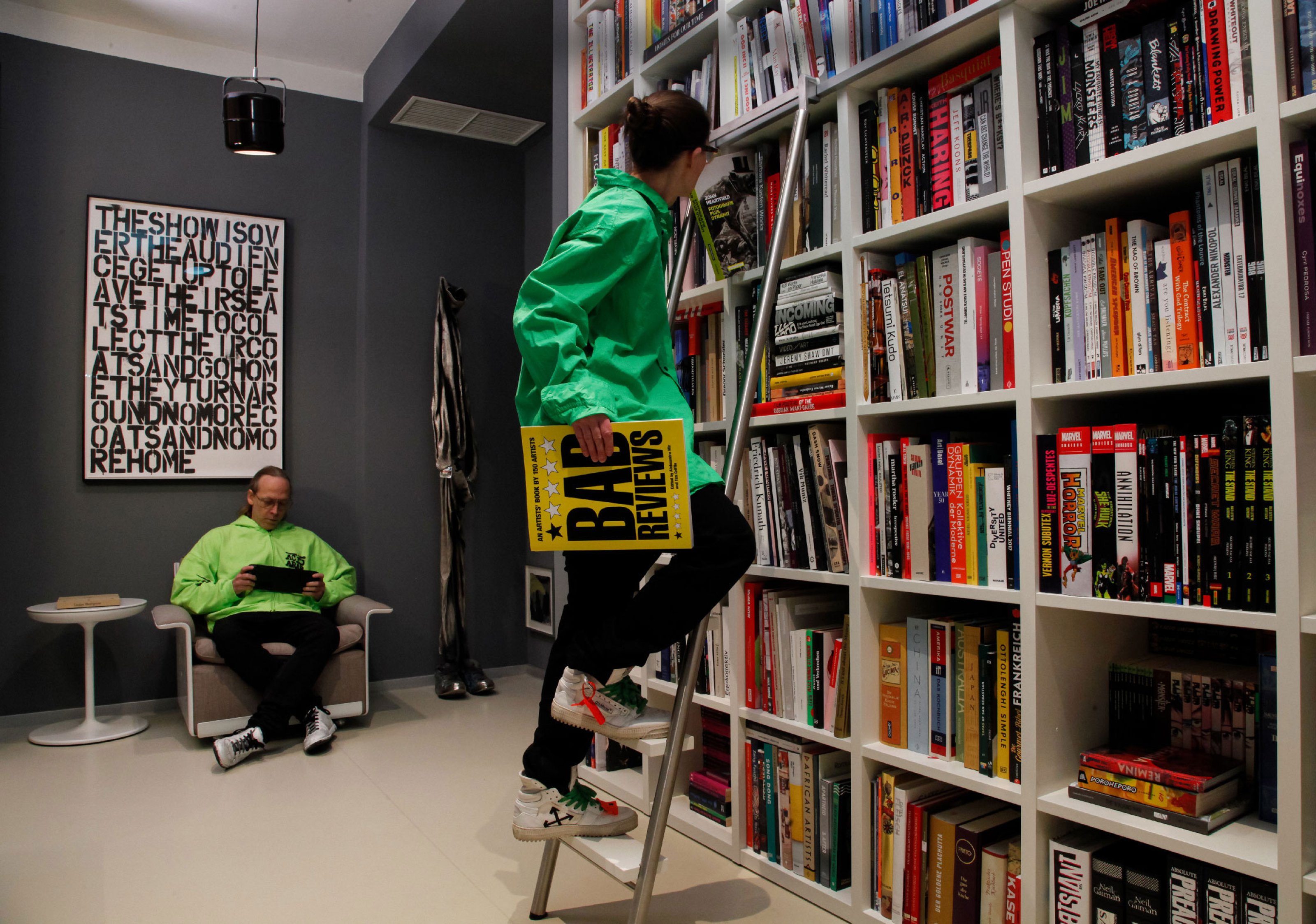 A woman in a green coat, climbing the white ladder against a library full of books, a man in a green coat sitting in the back seat.