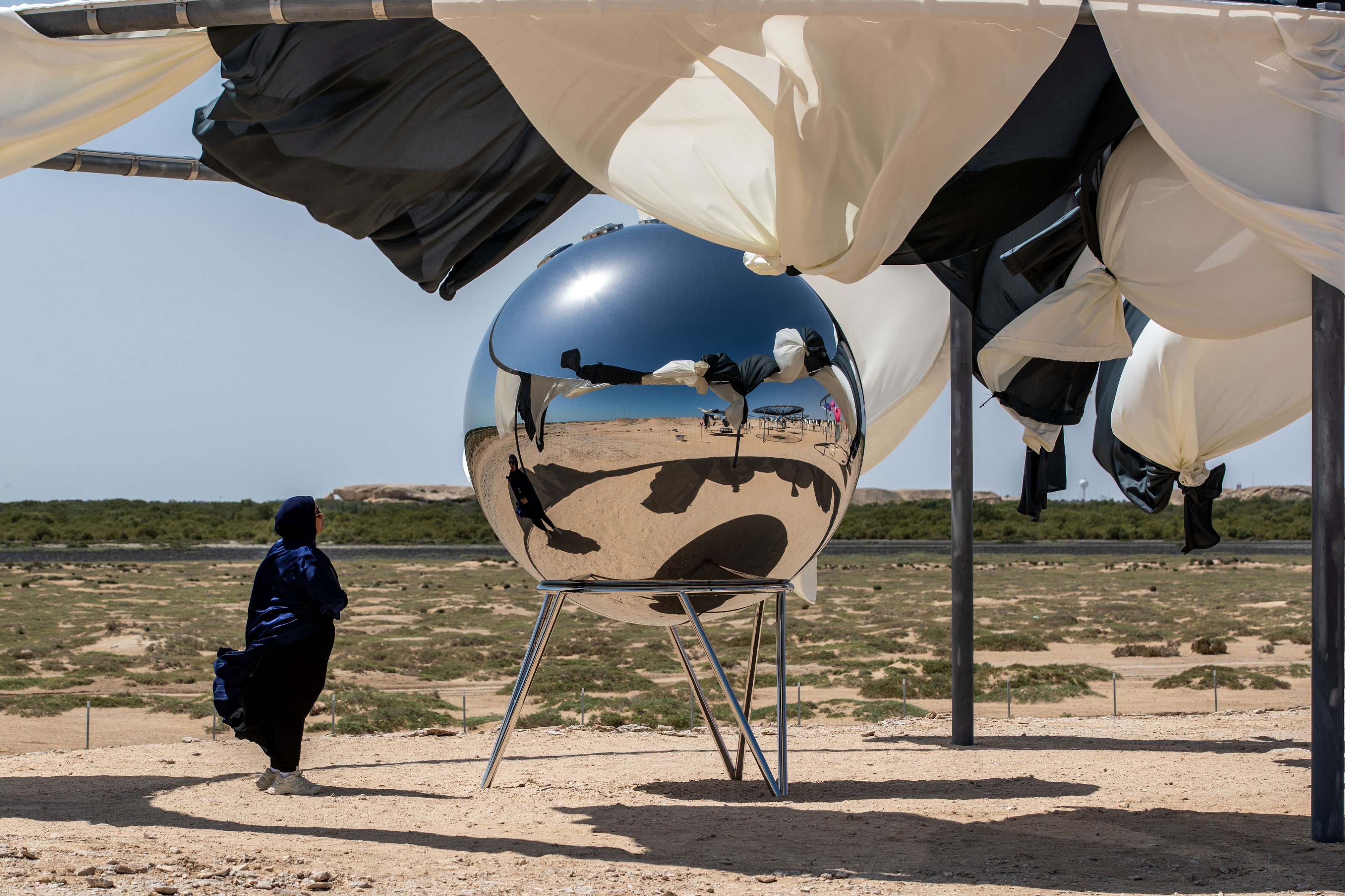 A woman in a black scarf staring at a metallic sphere set on metal legs in the middle of a desert.