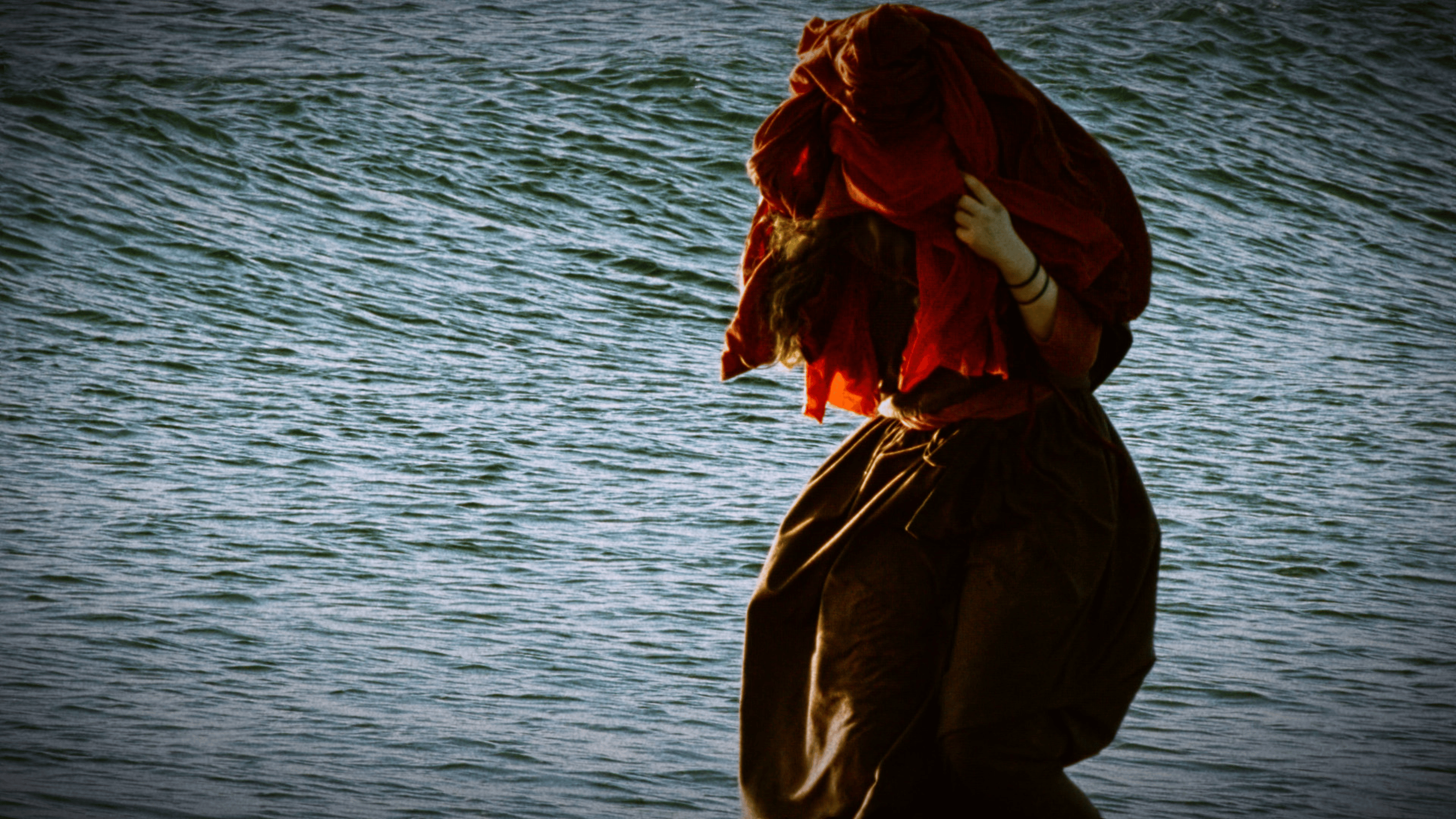 A woman standing with the sea behind her, completely covering her head with her clothes, in a red loose-fitting outfit.