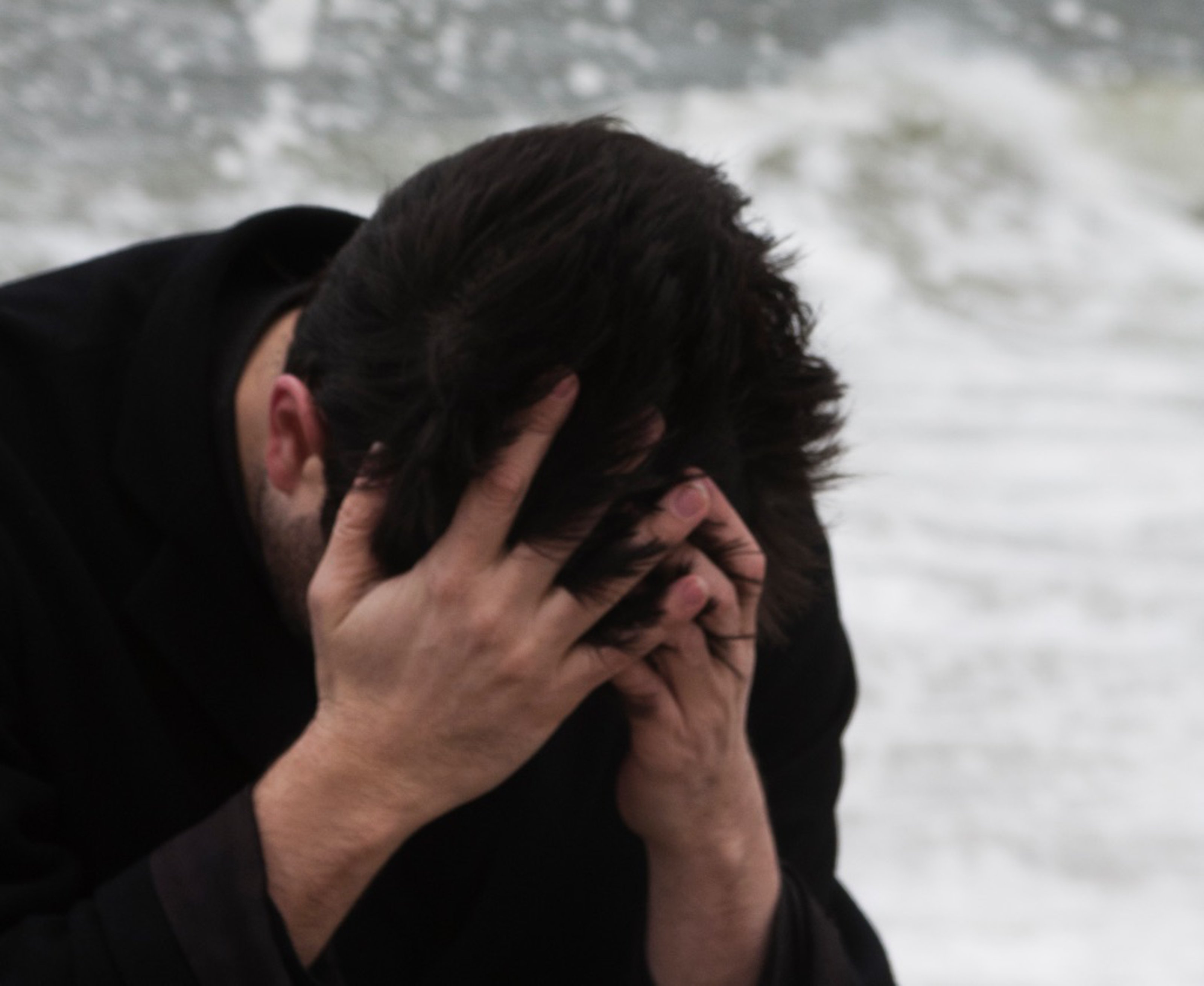 David Horvitz with a black sweater and his head in his hands standing in front of waves crashing.