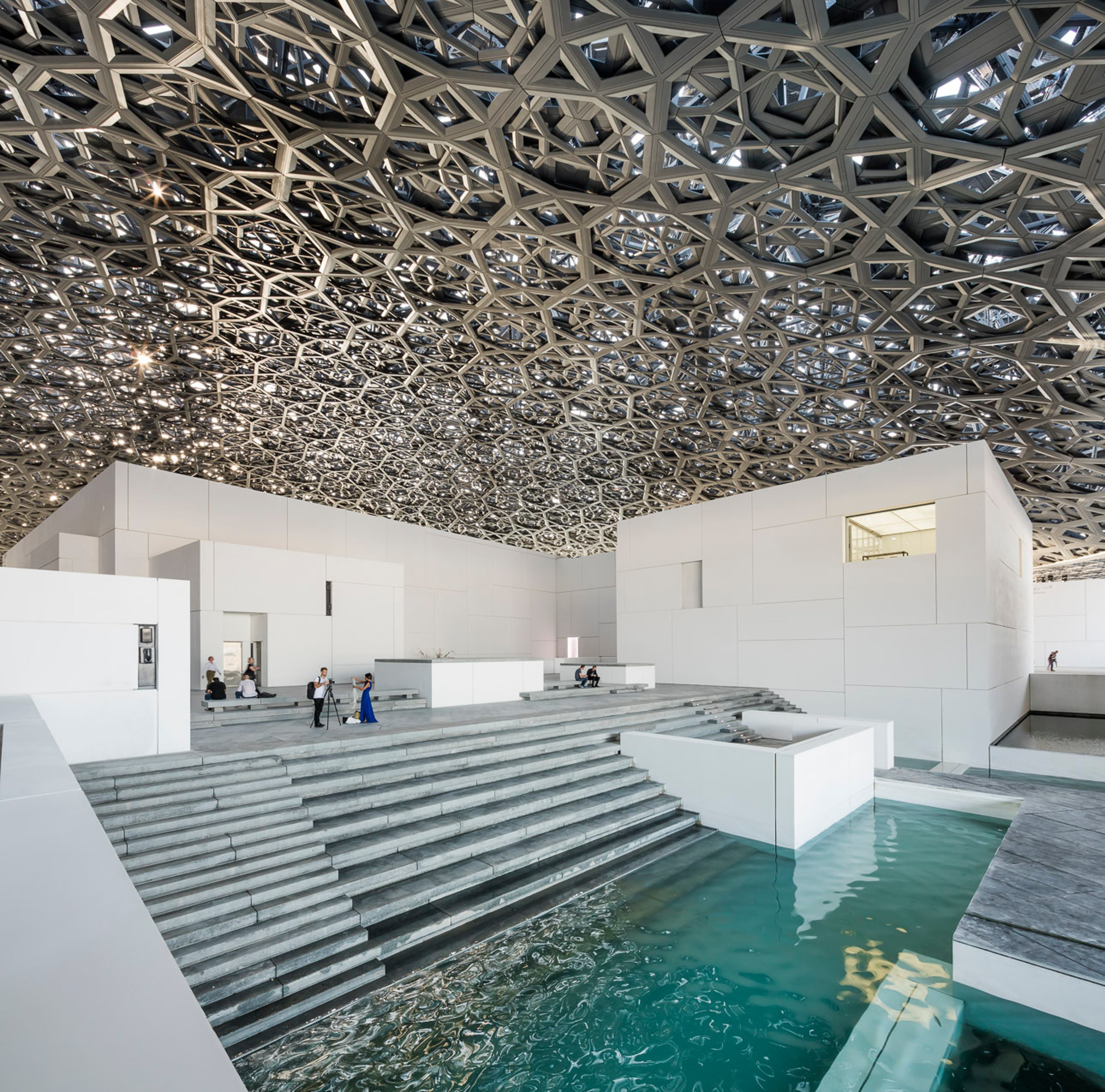 Interior view of Louvre Abu Dhabi with high and wide stairways, white geometric decorations on the ceiling and a pond inside.