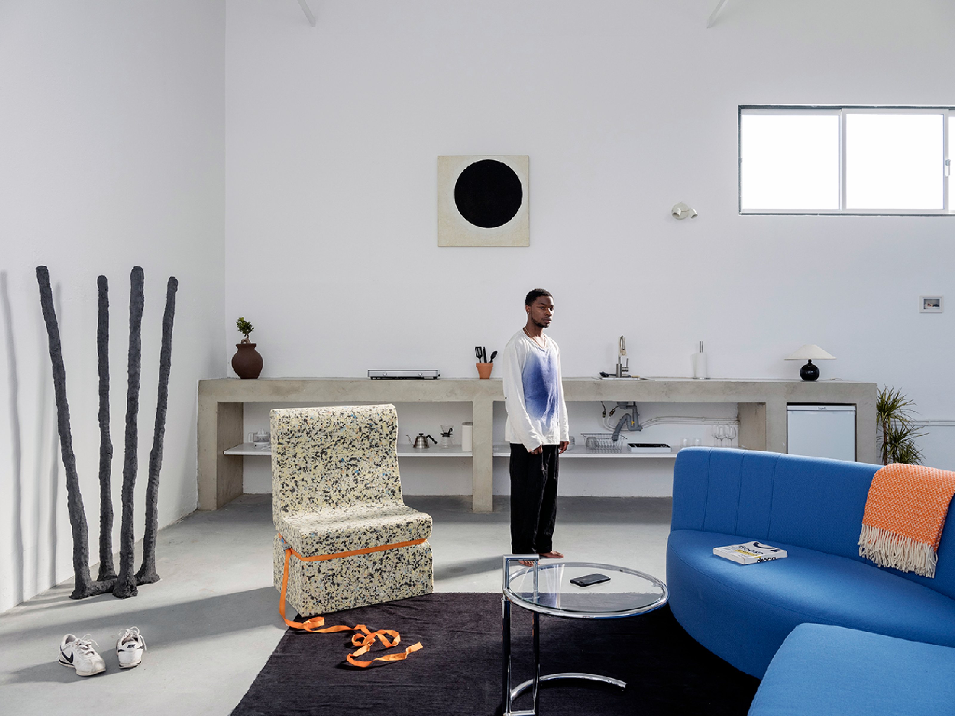 Dozie Kanu standing in a high ceiling space with white walls, minimal beige and gray colored objects around, a blue armchair and a black rug.