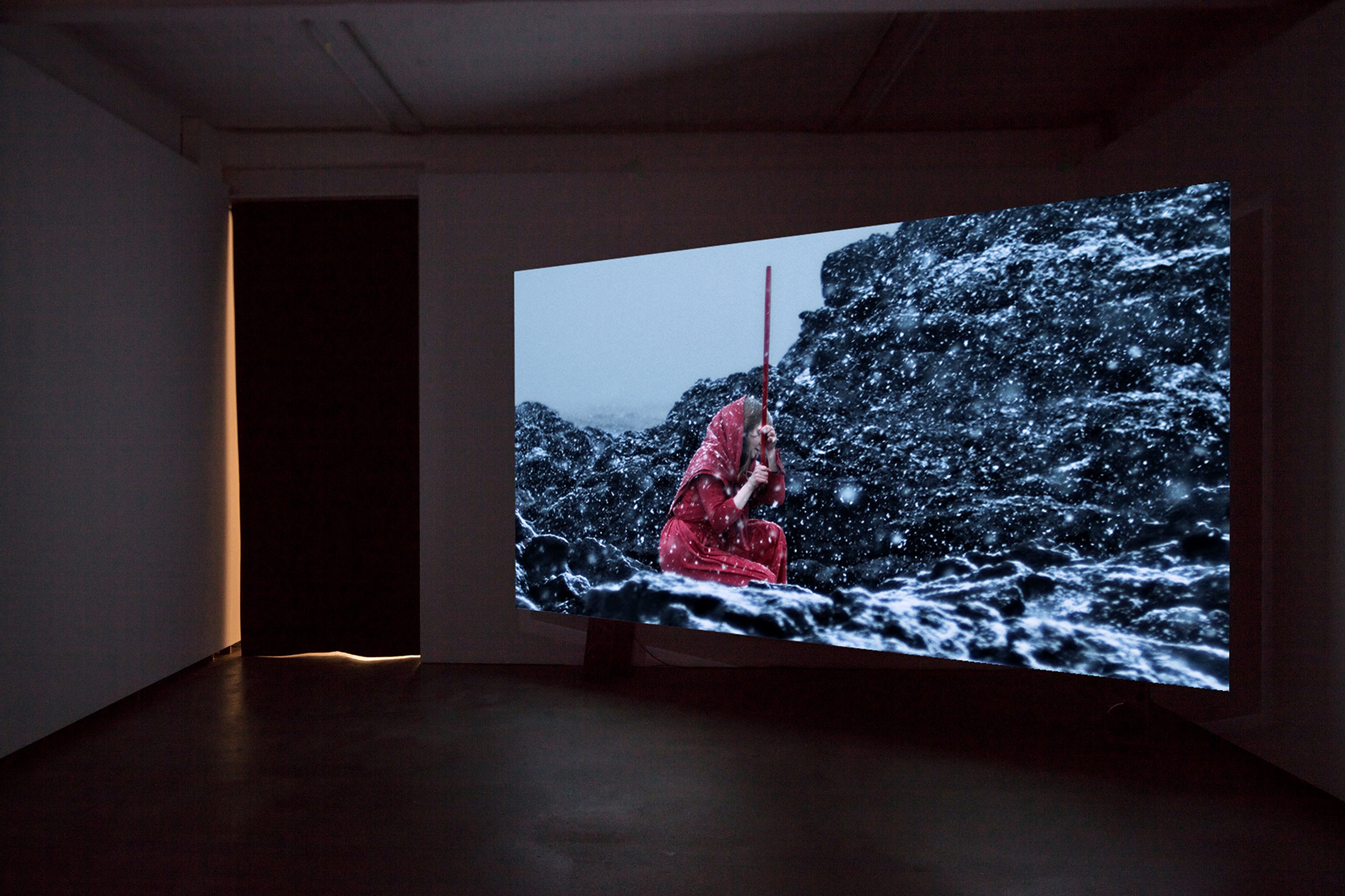 Video installed in a gallery space, on the projection is a woman sitting on a rocky surface, holding onto a long stick. There is snowfall around her