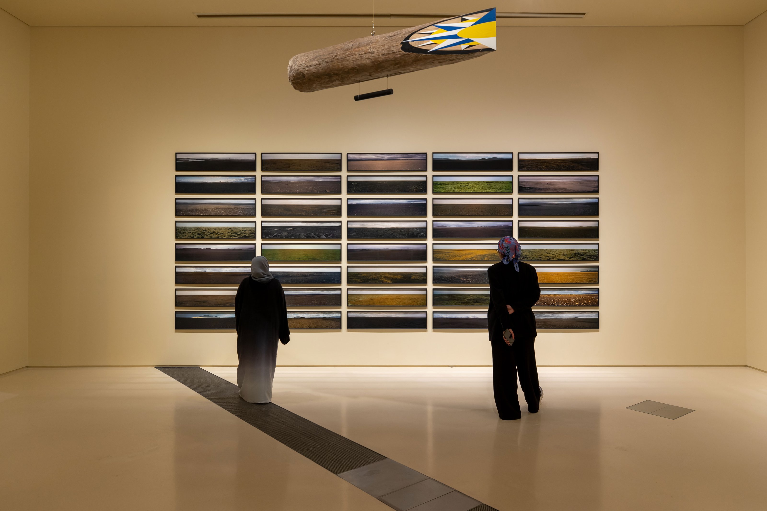 2 people looking at 40 rectangular paintings arranged neatly on the off-white wall of a high-ceilinged art gallery.