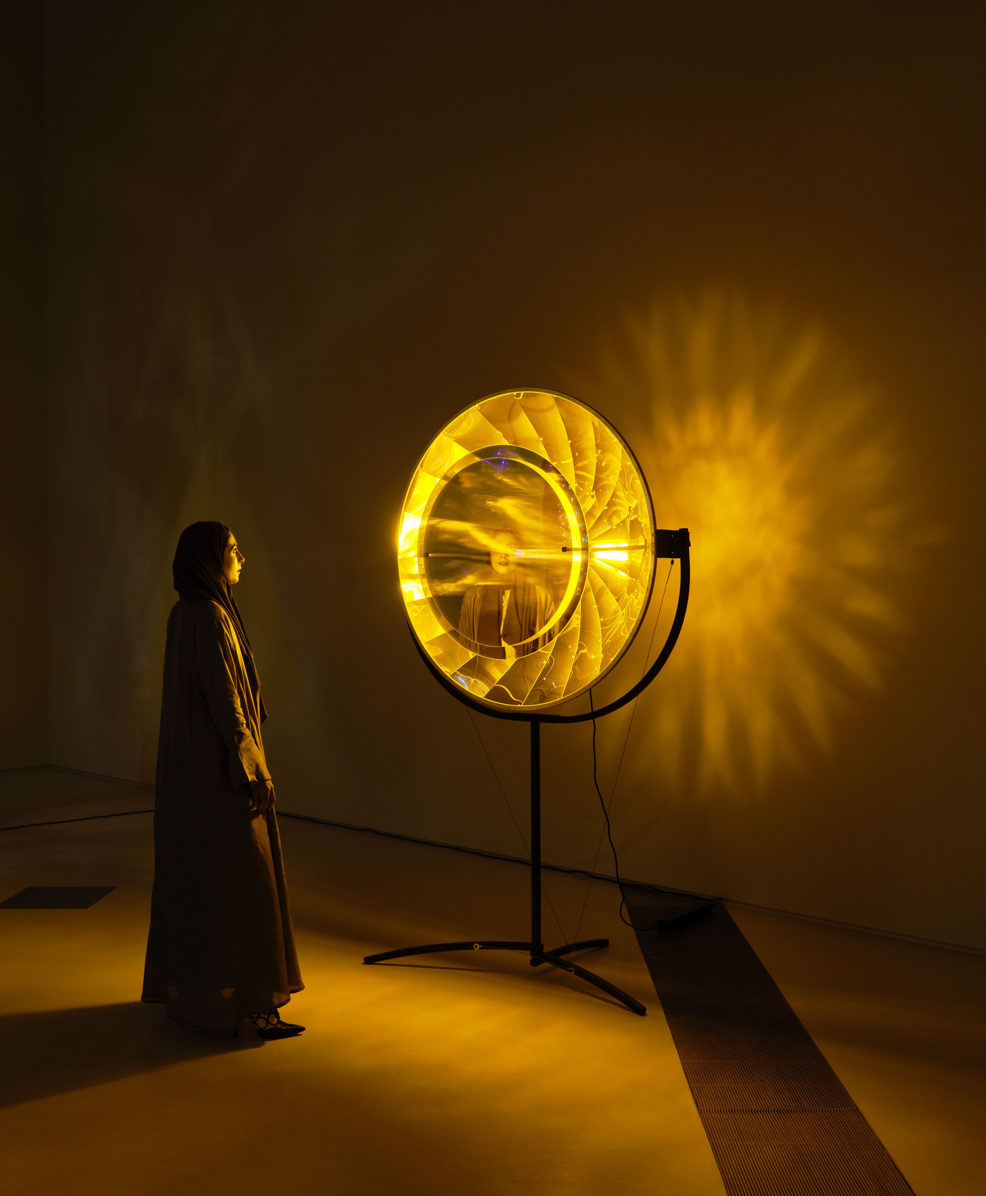 A woman in a scarf stares at a large transparent, circular stone, set on metal legs, illuminated by yellow light.