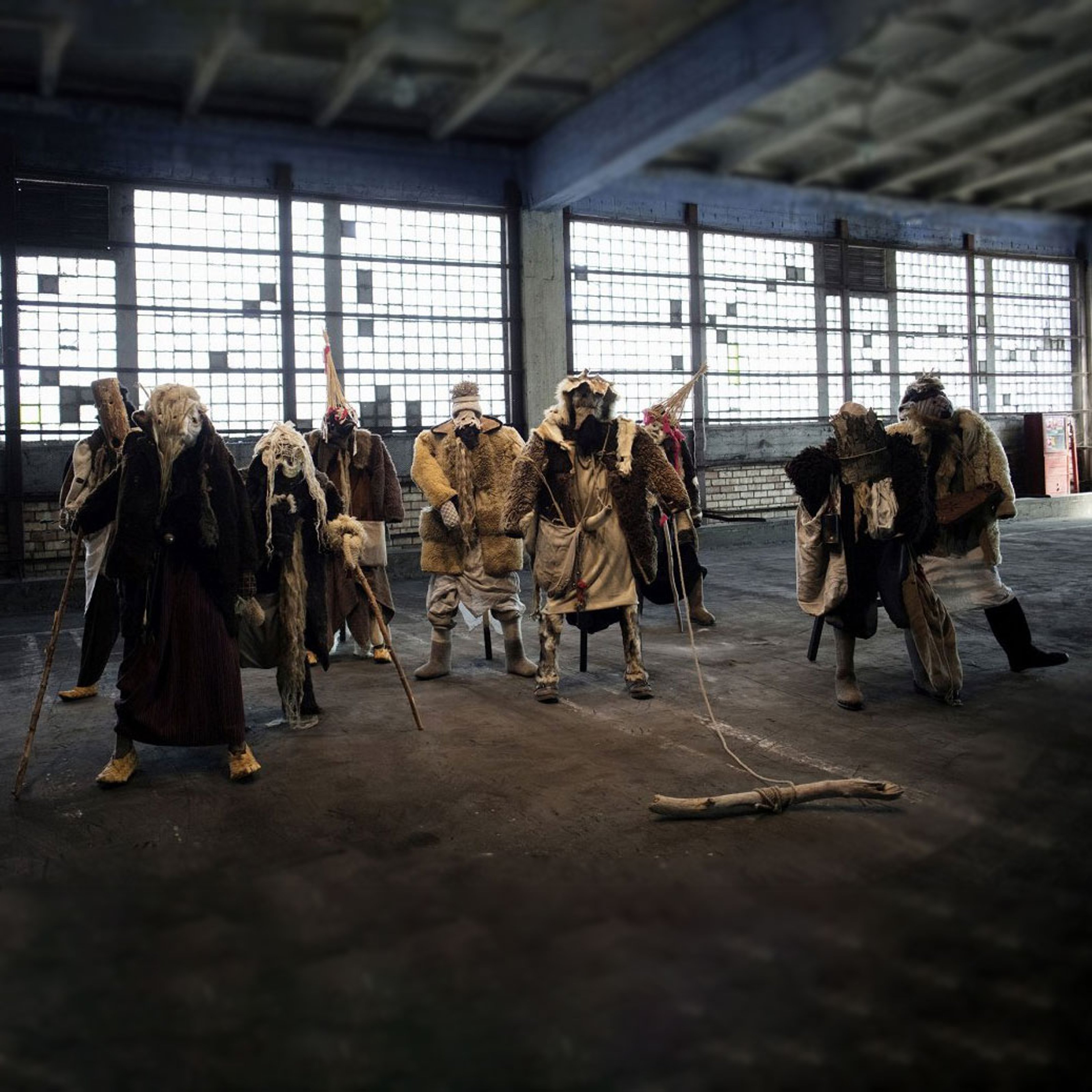 9 old and layered dressed mannequins standing in an old concrete space with large windows.