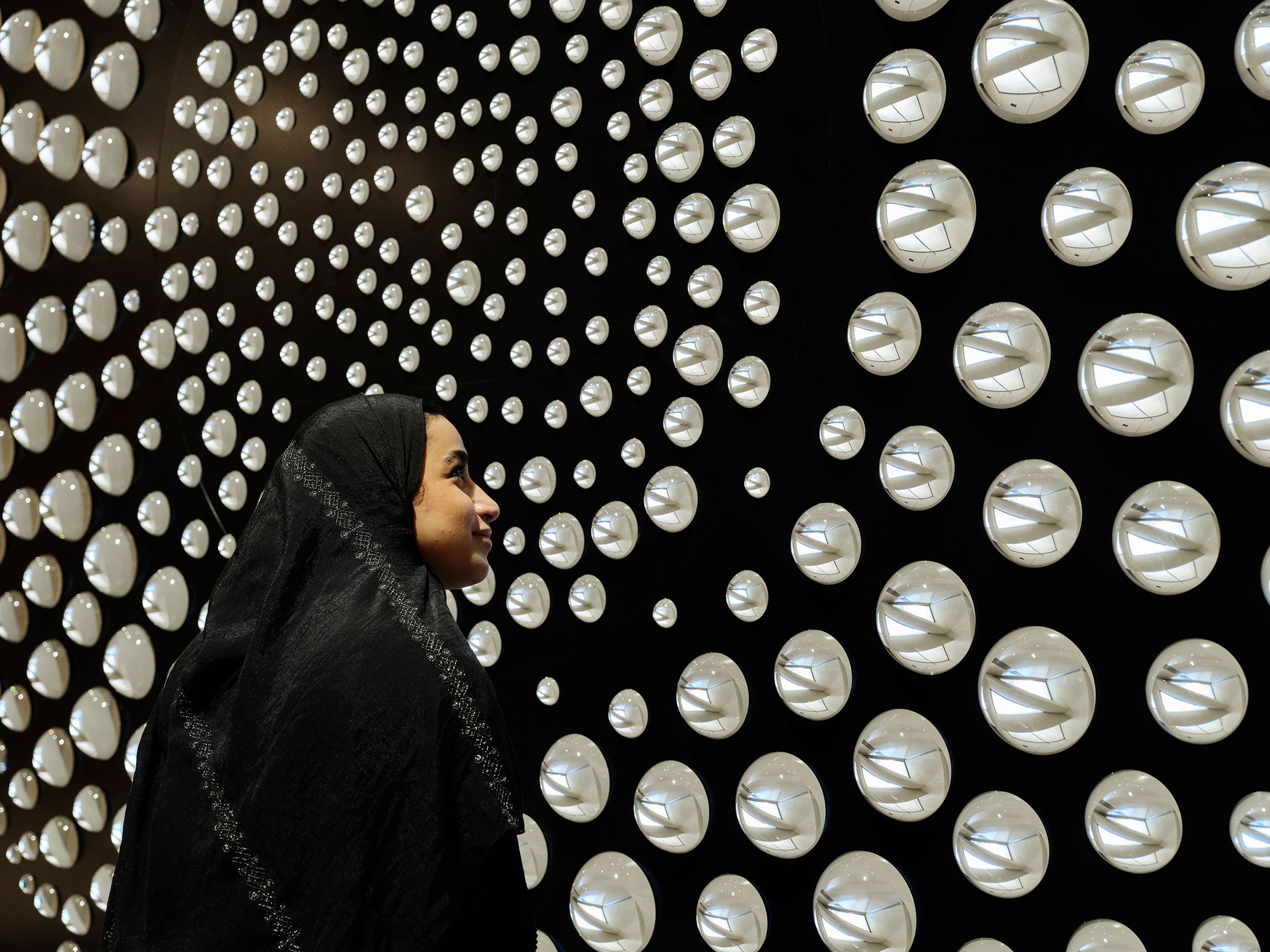 A woman in a black scarf is looking at a black wall over which small, round, shiny lenses hang.