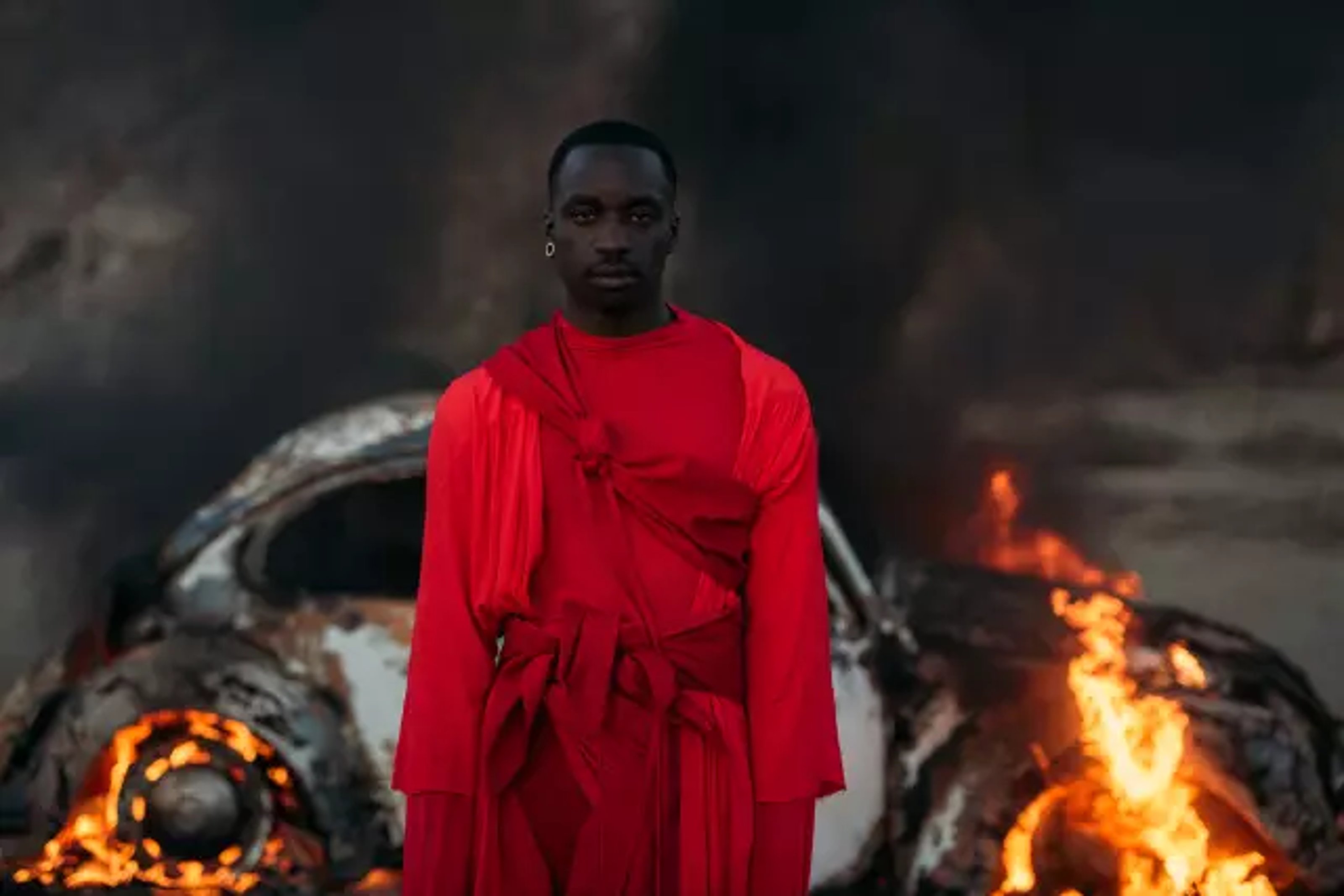 A man wrapped in loose red fabric standing in front of a burning white car.