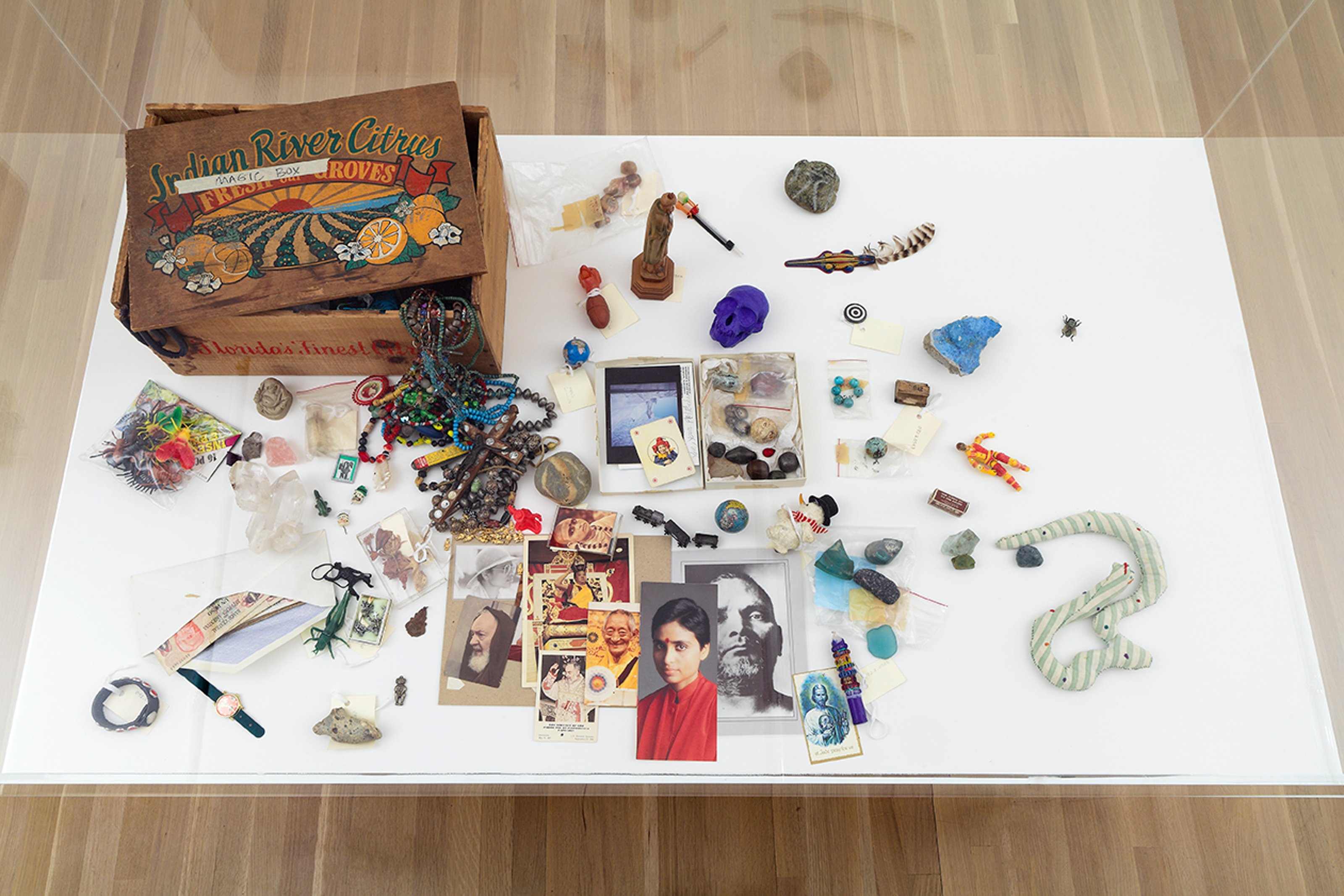 A cardboard box on the floor of a larger box with a white bottom and transparent sides, containing colorful objects, beaded jewelry, and photographs.