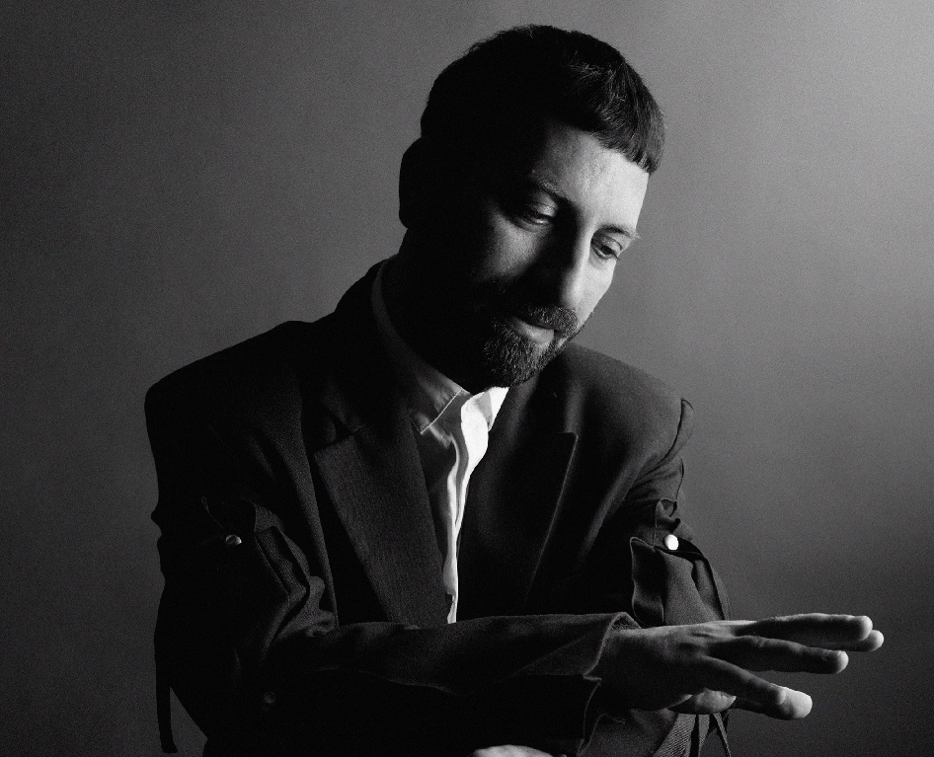 A contrast black-and-white portrait photograph of Ahmet Öğüt sitting on a chair and looking at his hand.