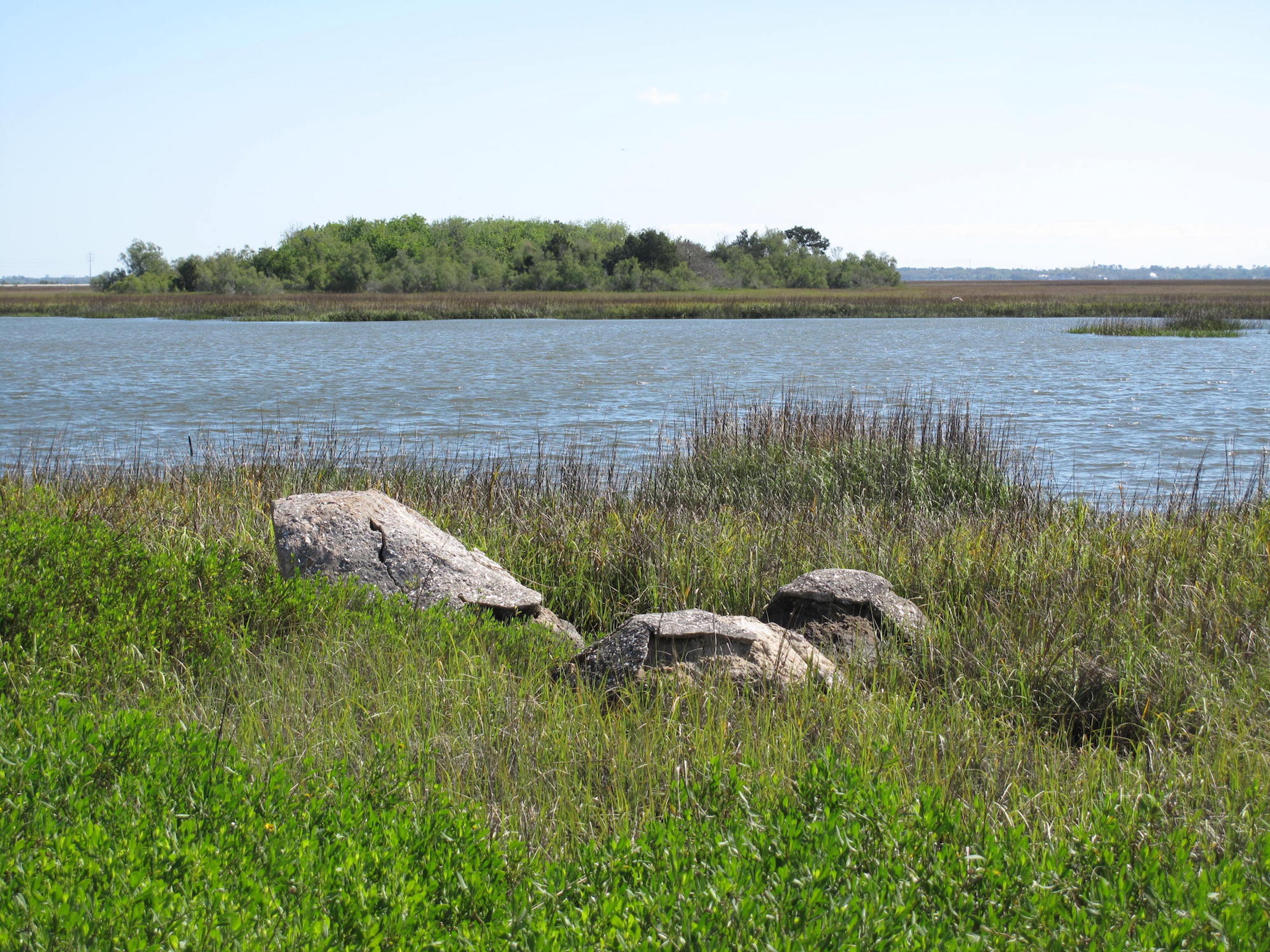Three rocks in a grassy landscape beside a tranquil lake, with a scenic view of the opposite shore in daylight.