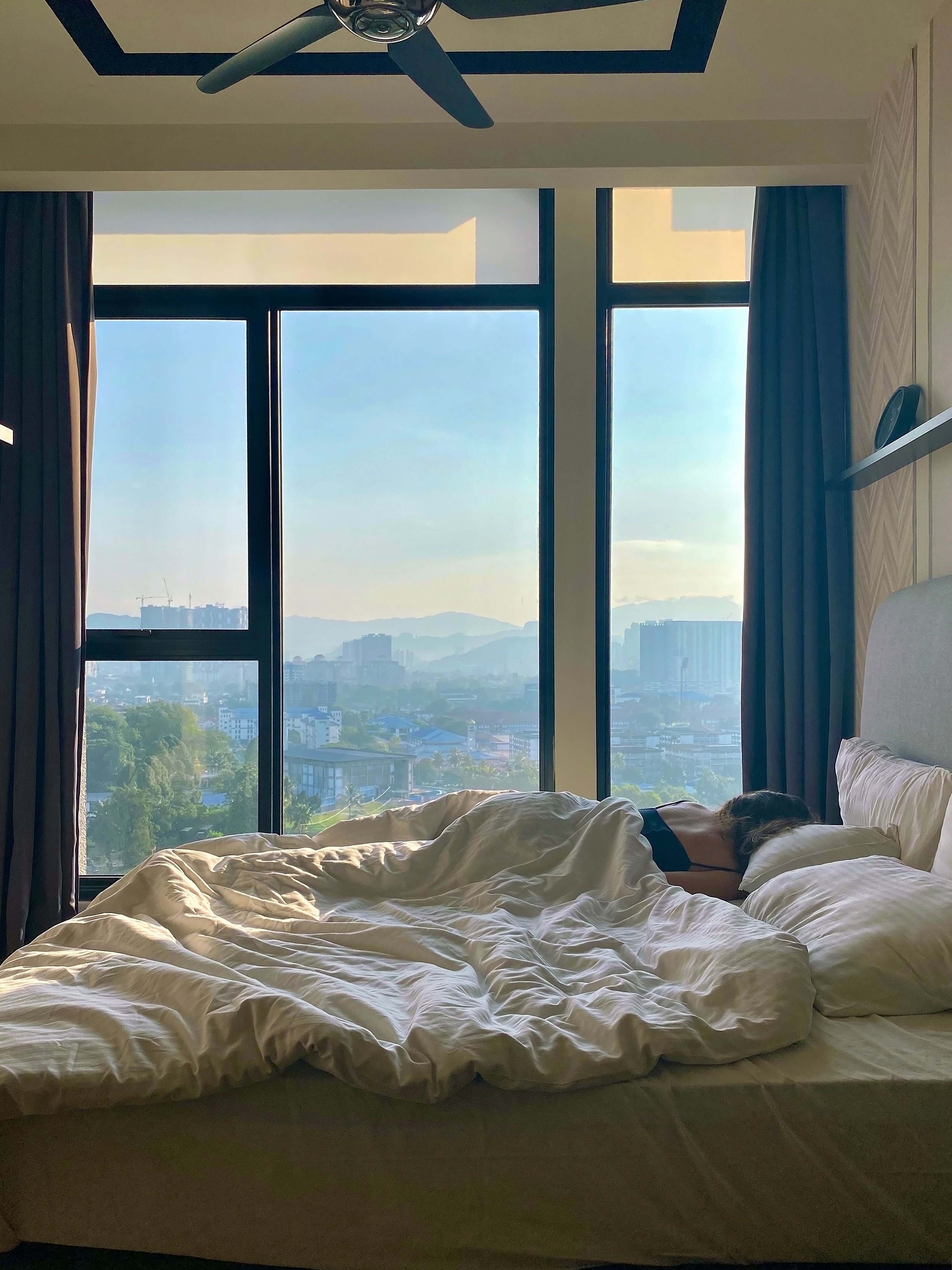 A woman sleeping with her back on the bed in front of a large window during the daytime.