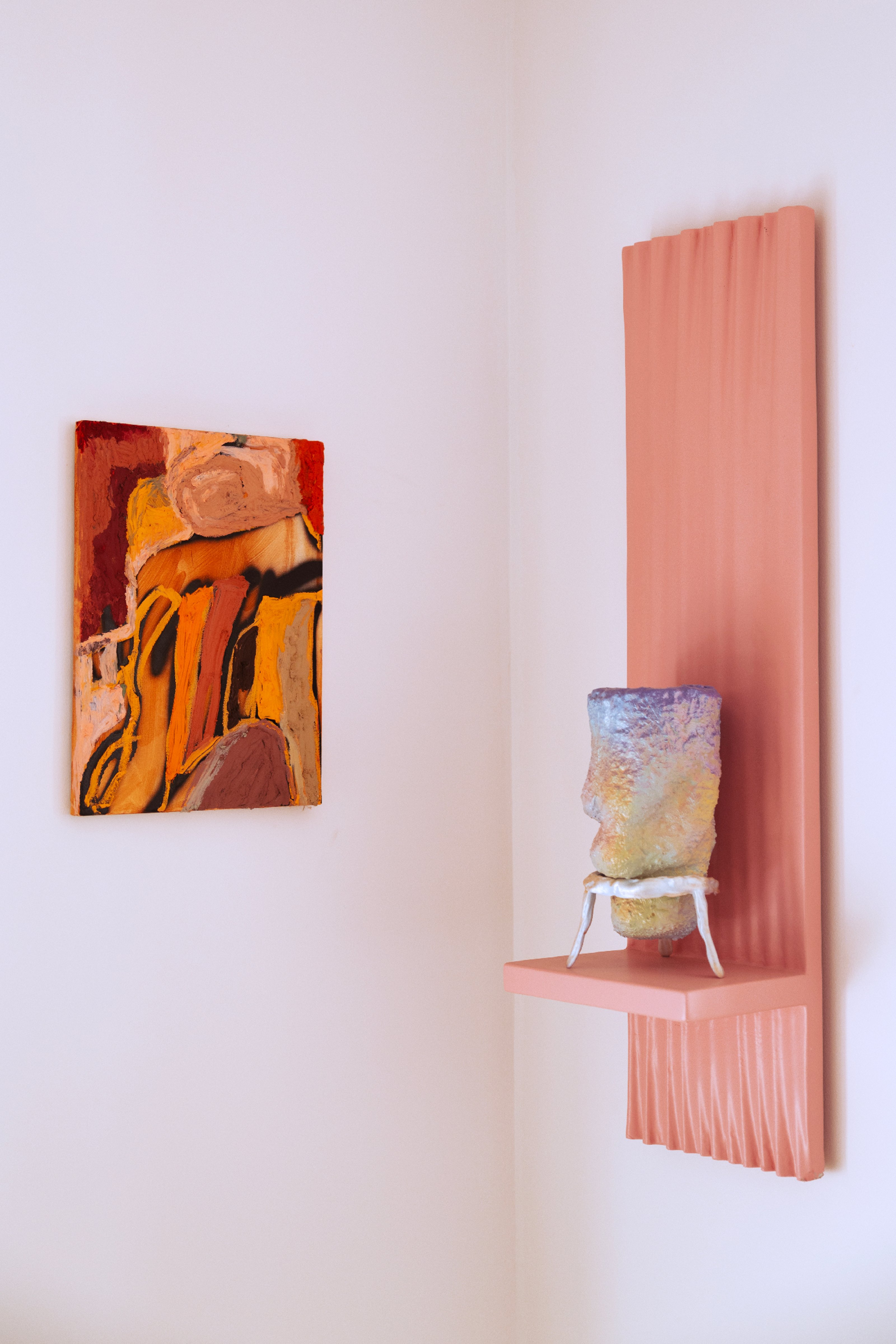 An abstract painting in shades of pink placed in the corner of a white-walled room. A ceramic object on a wall-mounted pink shelf opposite.