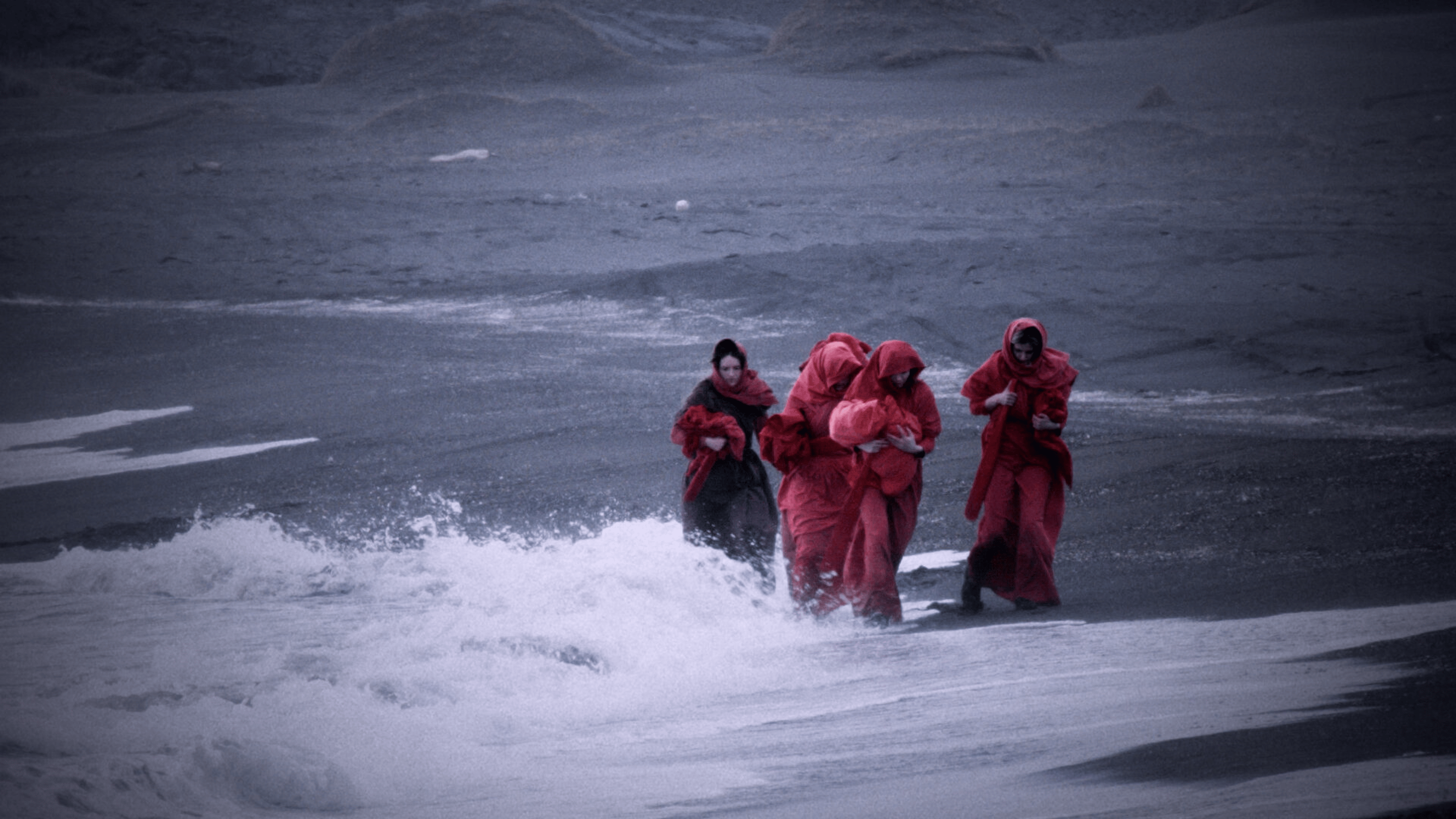 Women in red garbs walking by the sea, with waves hitting them.
