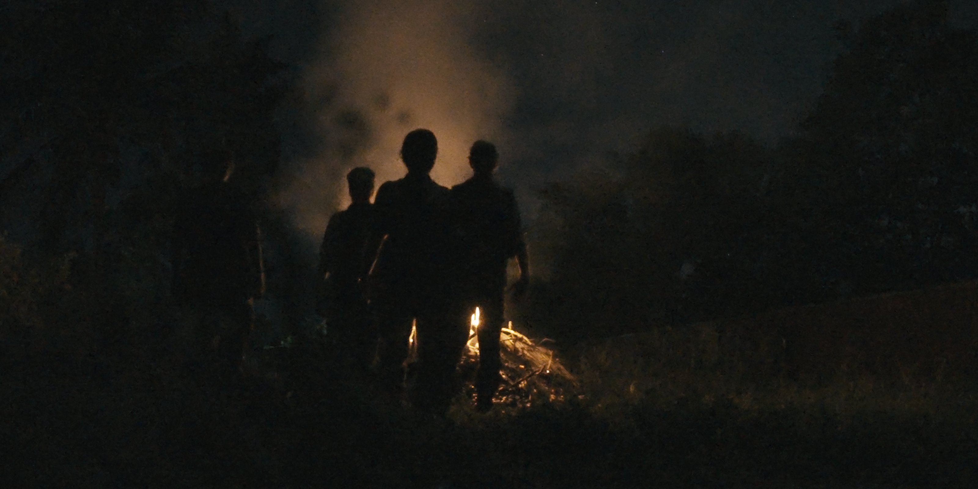 A silhouette of three people looking at a lit fire at dusk, seen from behind.