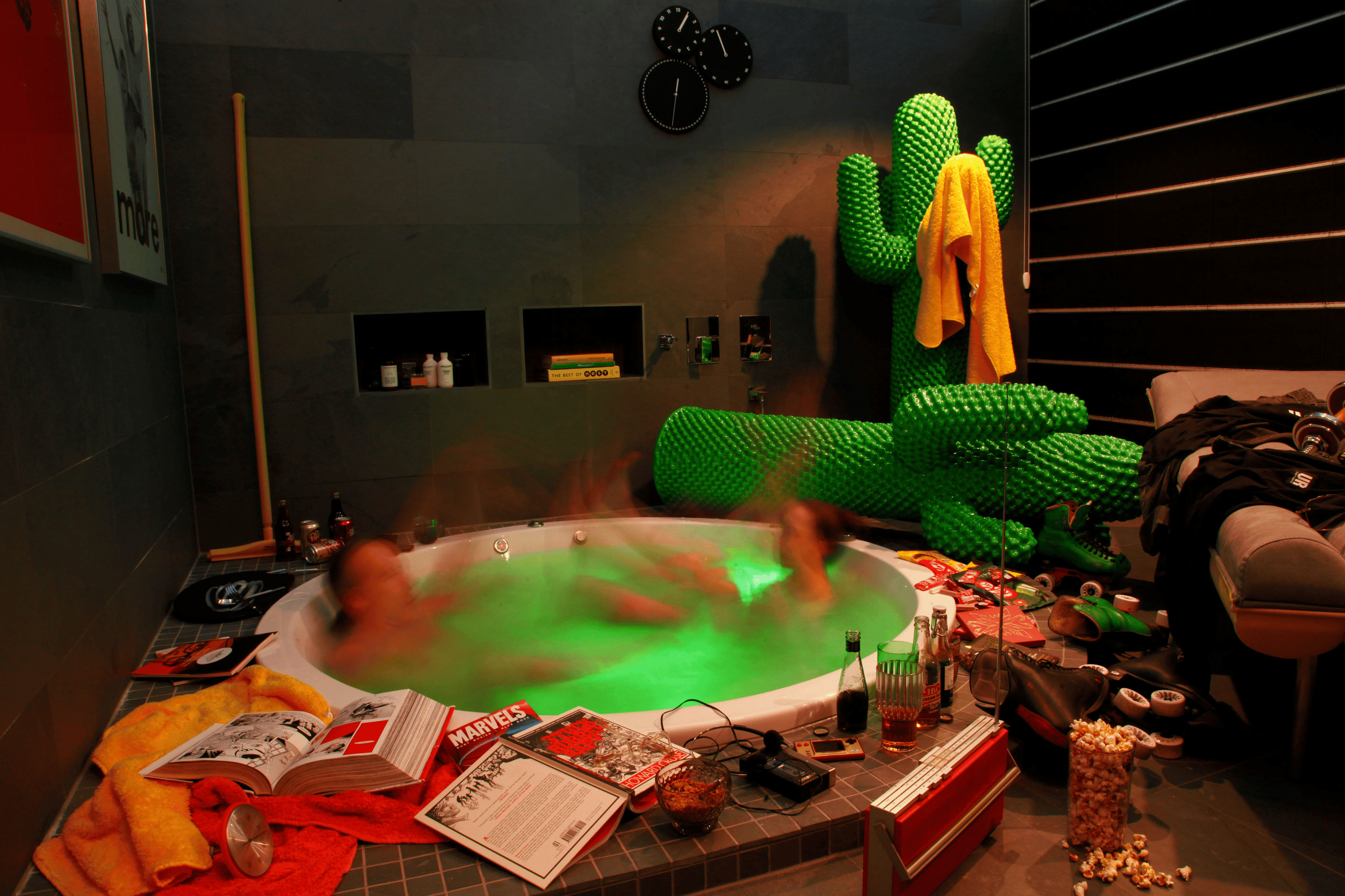 Two people, blurry, in jacuzzi. They're in a room with some disorganized objects, gigantic inflatable cacti and some magazines laying around.