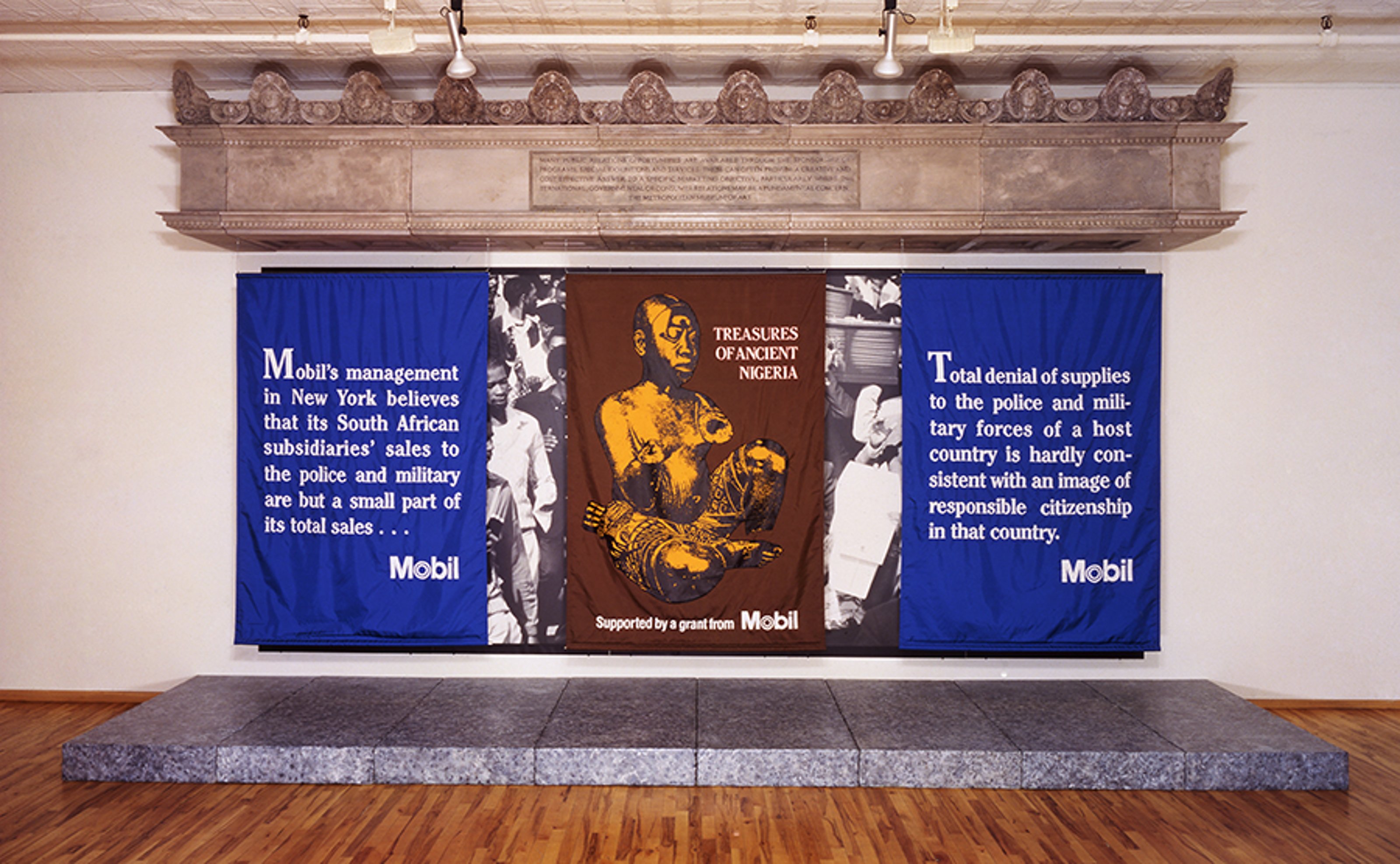 Haacke's installation. Three pieces of fabric hung like flags or a billboard ad. The installation contains statements from Mobil, the oil company. Descriptions from left to right: First one reads “ Mobil’s management in New York believes that its South African subsidiaries’ sales to the police and military are but a small part of its total sales…” In between is an image of a crowd, possibly from South Africa. The next fabric reads: “Treasures of Ancient Nigeria, Supported by a grant from Mobil.” With an image of a traditional Nigerian sculpture of a human form. In between is an image again from a crowd. The final fabric reads: “Total denial of supplies to the police and military forces of a host country is hardly consistent with an image of responsible citizenship in that country. Mobil.”