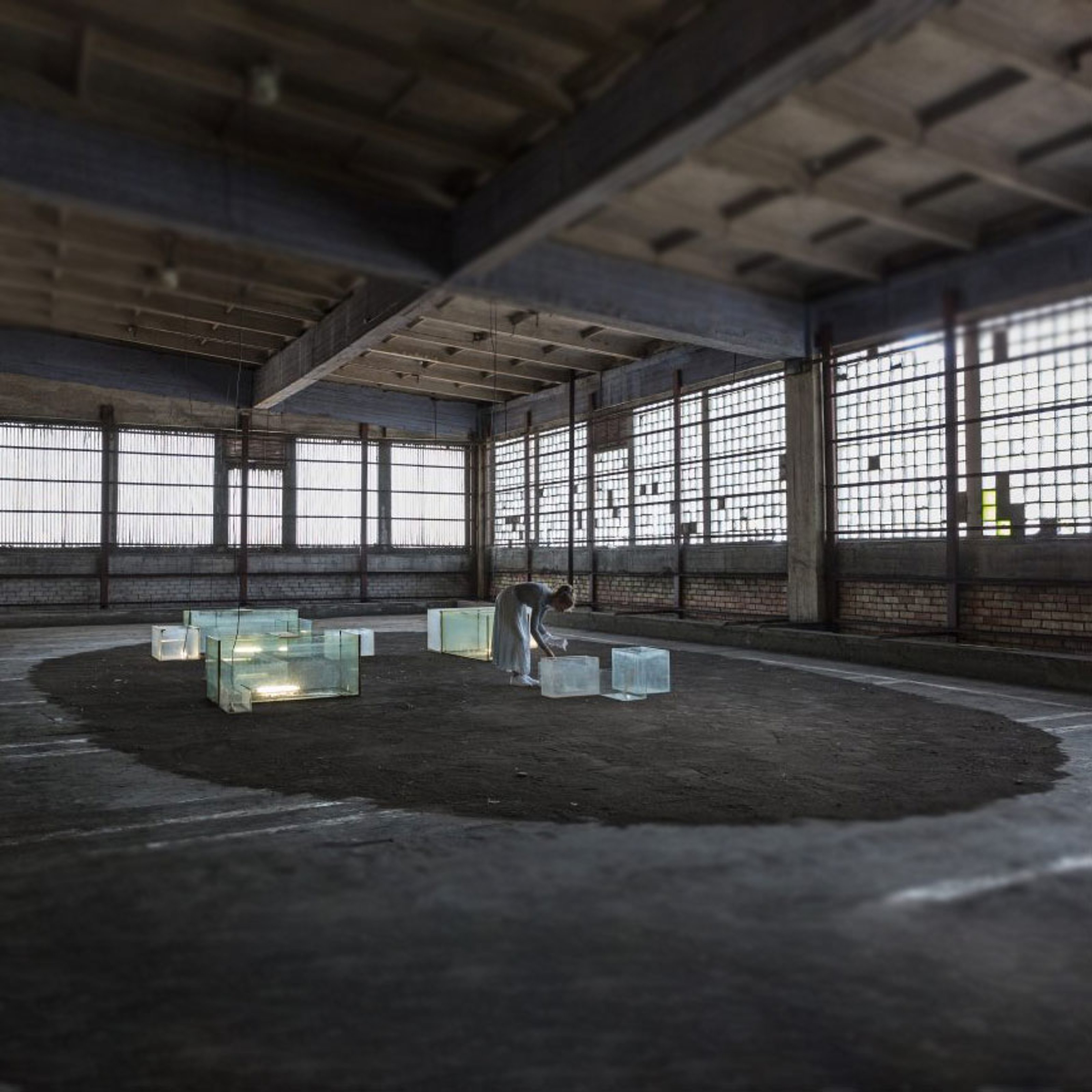 A woman in a white dress is bent over the rectangular glass prisms set on the floor of an old concrete room with large windows.