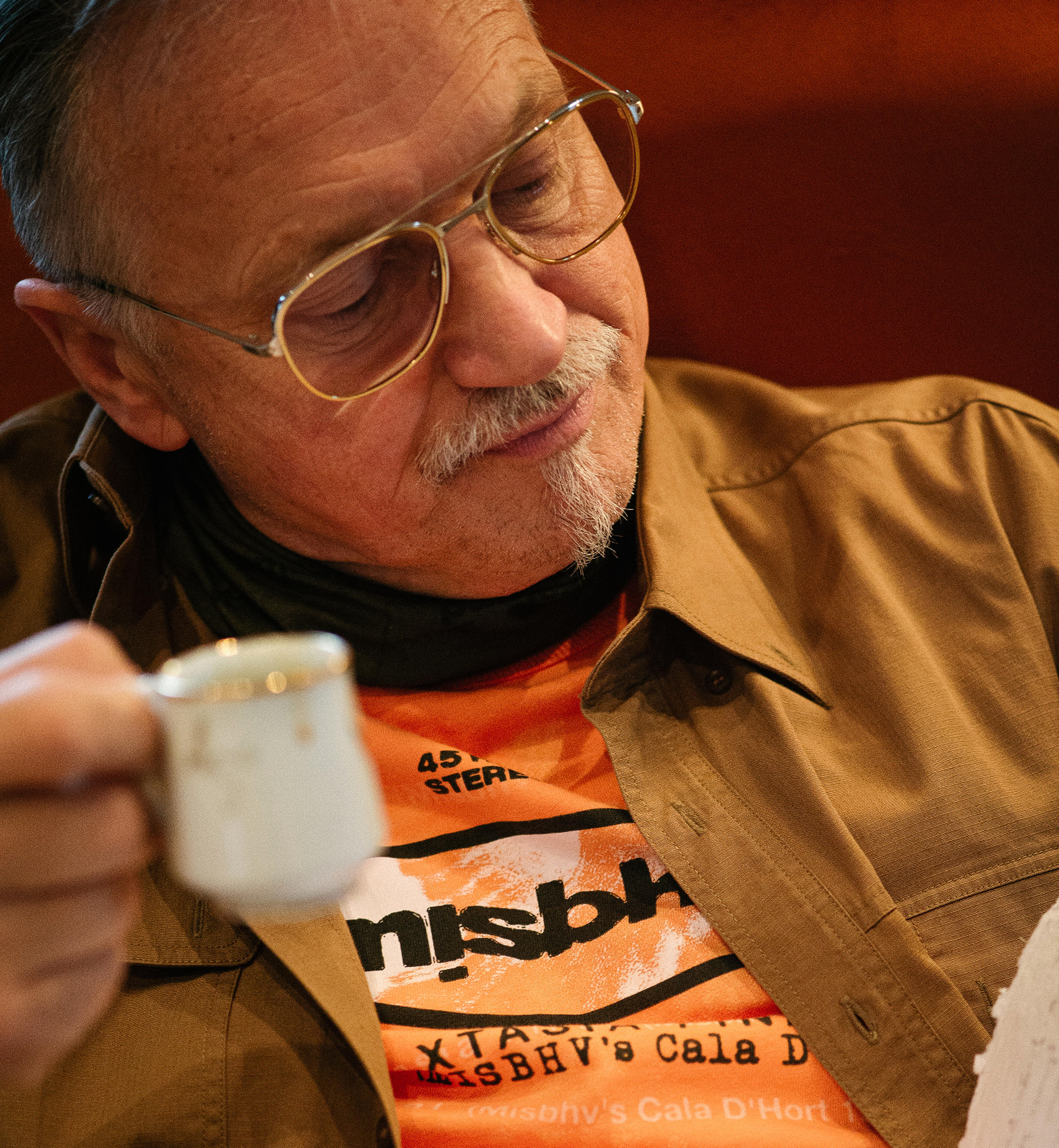 Janis Zuzans is reading a book, wearing glasses, an orange shirt and an ocher shirt, holding a small white coffee mug.