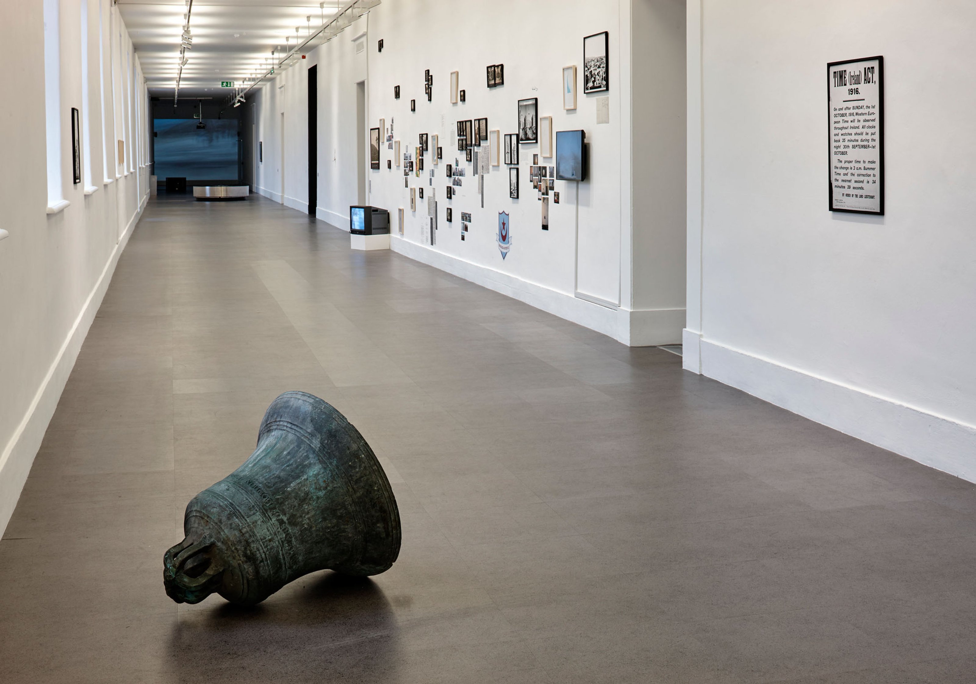 Various artworks showcased on a white gallery wall, including a big metal bell displayed in a long gray corridor.