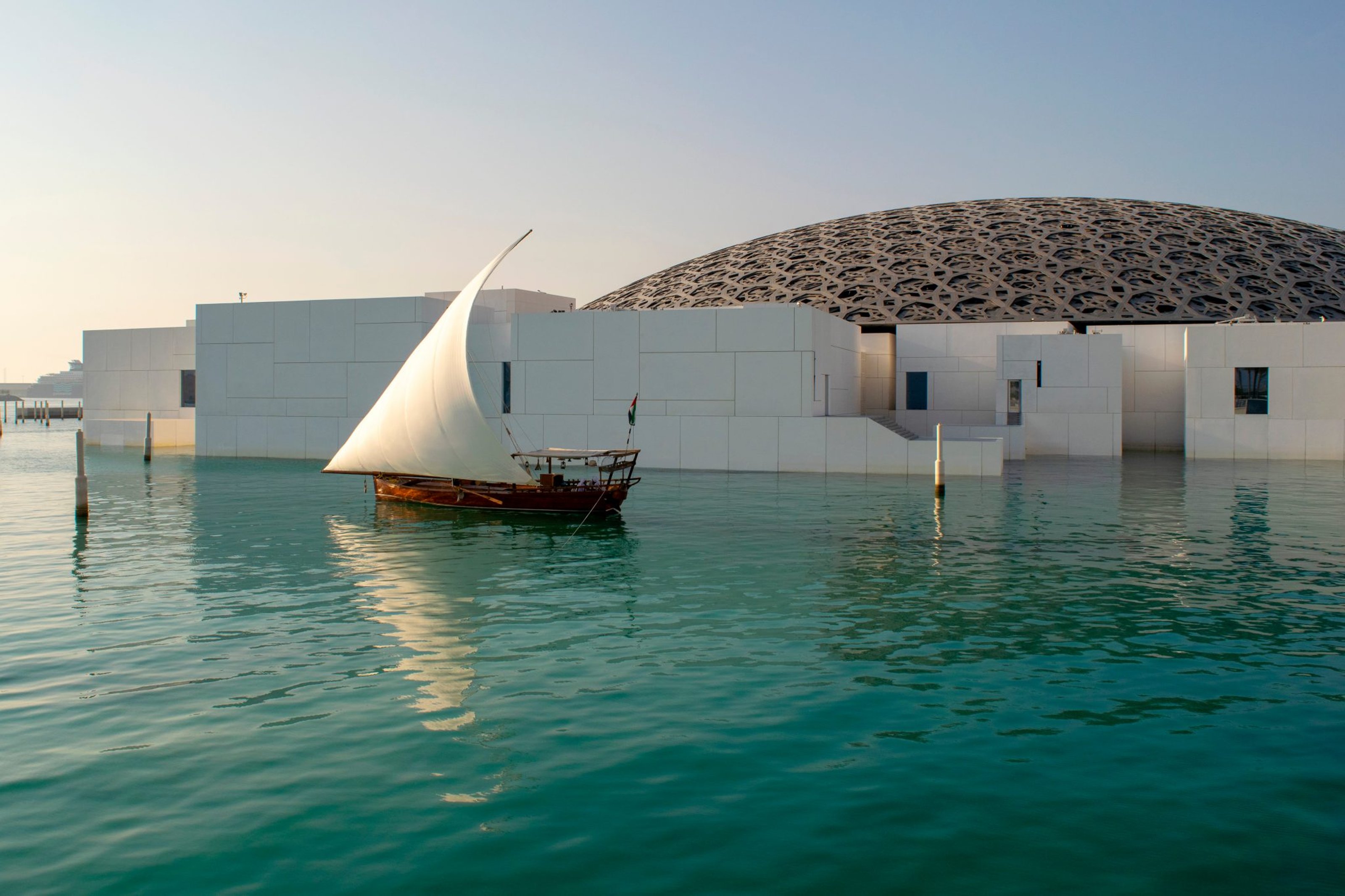 Outside view of Louvre Abu Dhabi, with a lake in front and a white sailboat on the water.