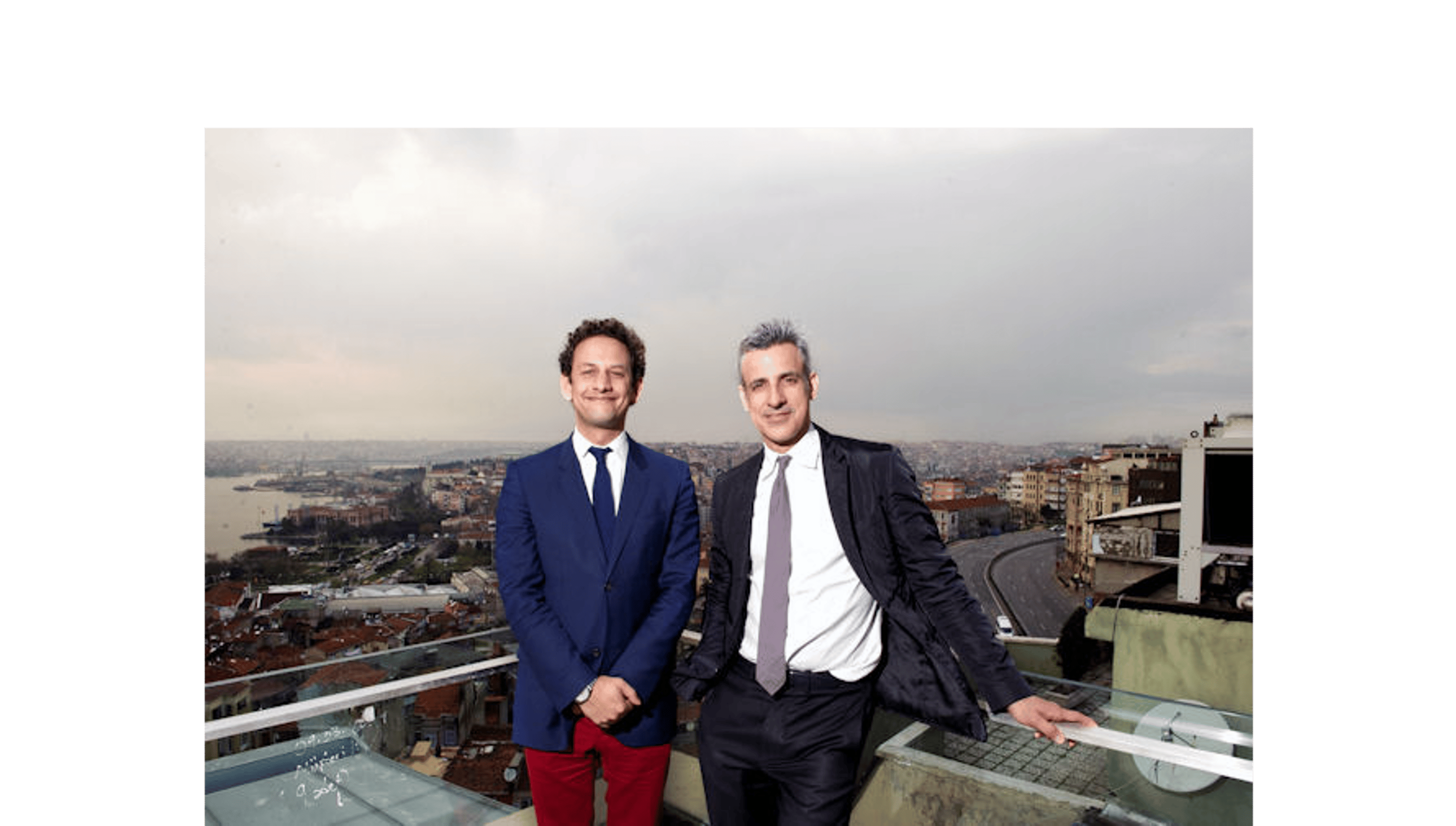 Two men in suits standing on the roof of a building with a view of the city behind them.
