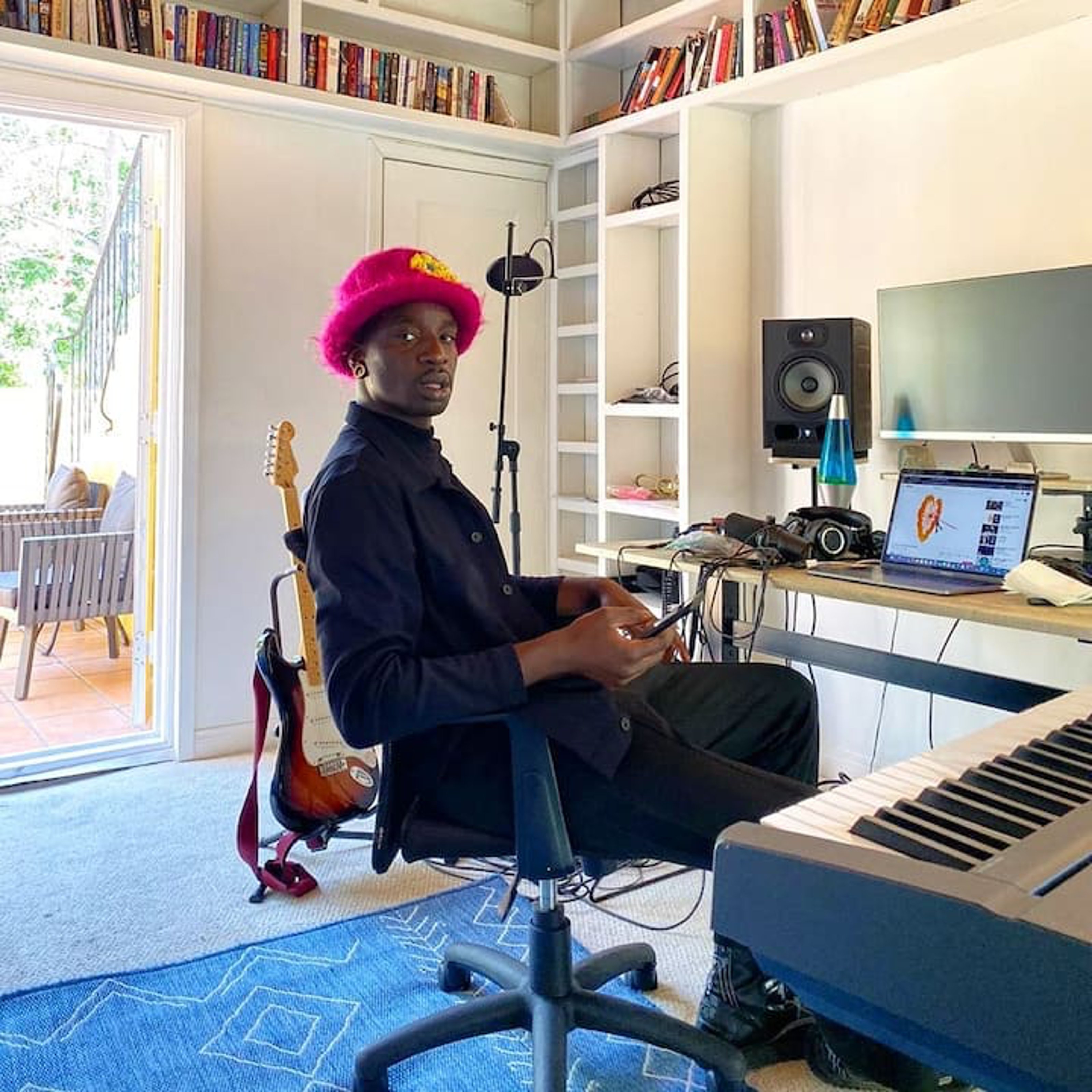 Sitting in a black chair, facing an organ, is a man dressed in black, wearing a pink feathered hat.