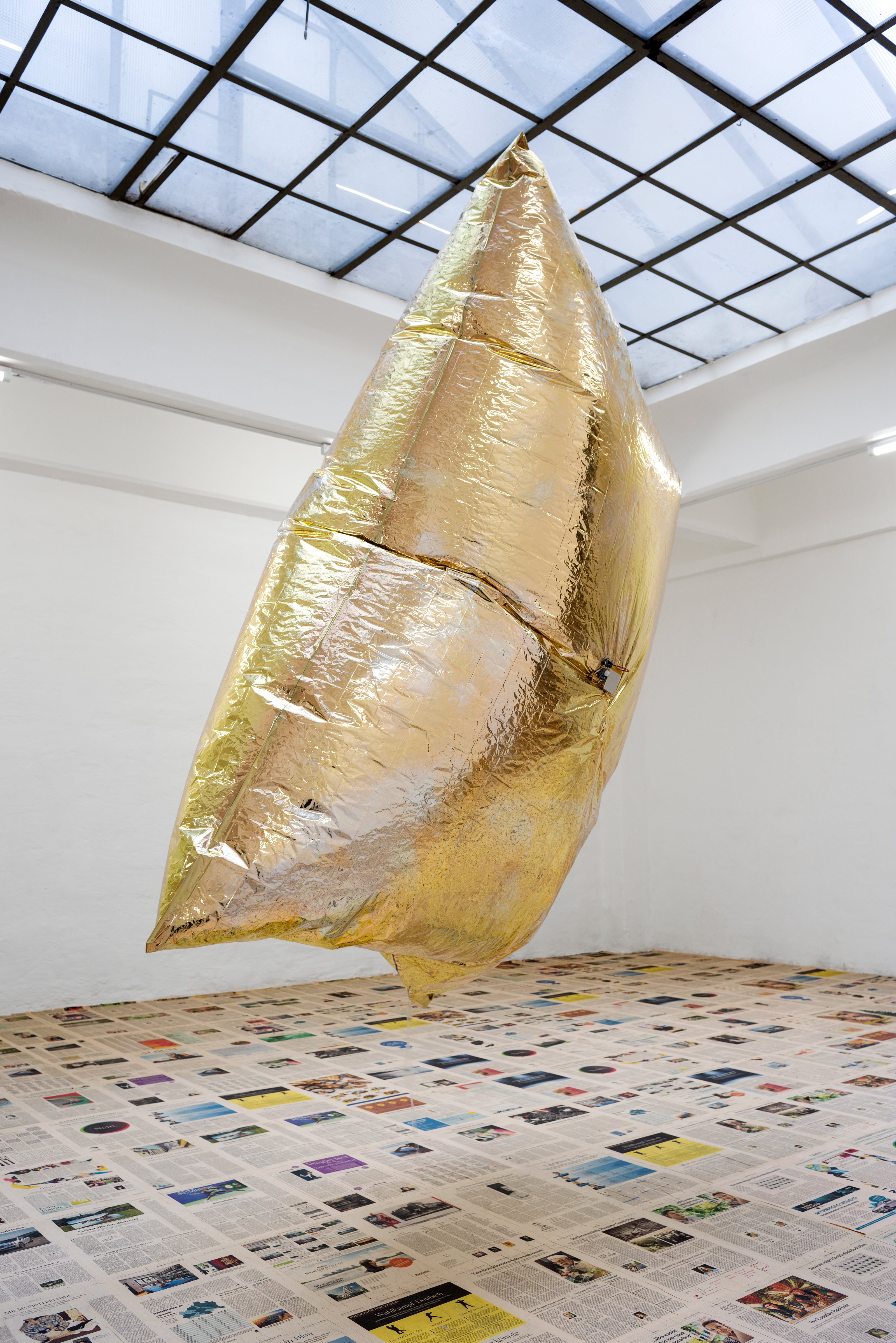 A huge, air-filled, golden, pillow-like balloon is suspended from the ceiling of an art gallery with a glass ceiling, white walls, and a floor covered with newspaper leaves.