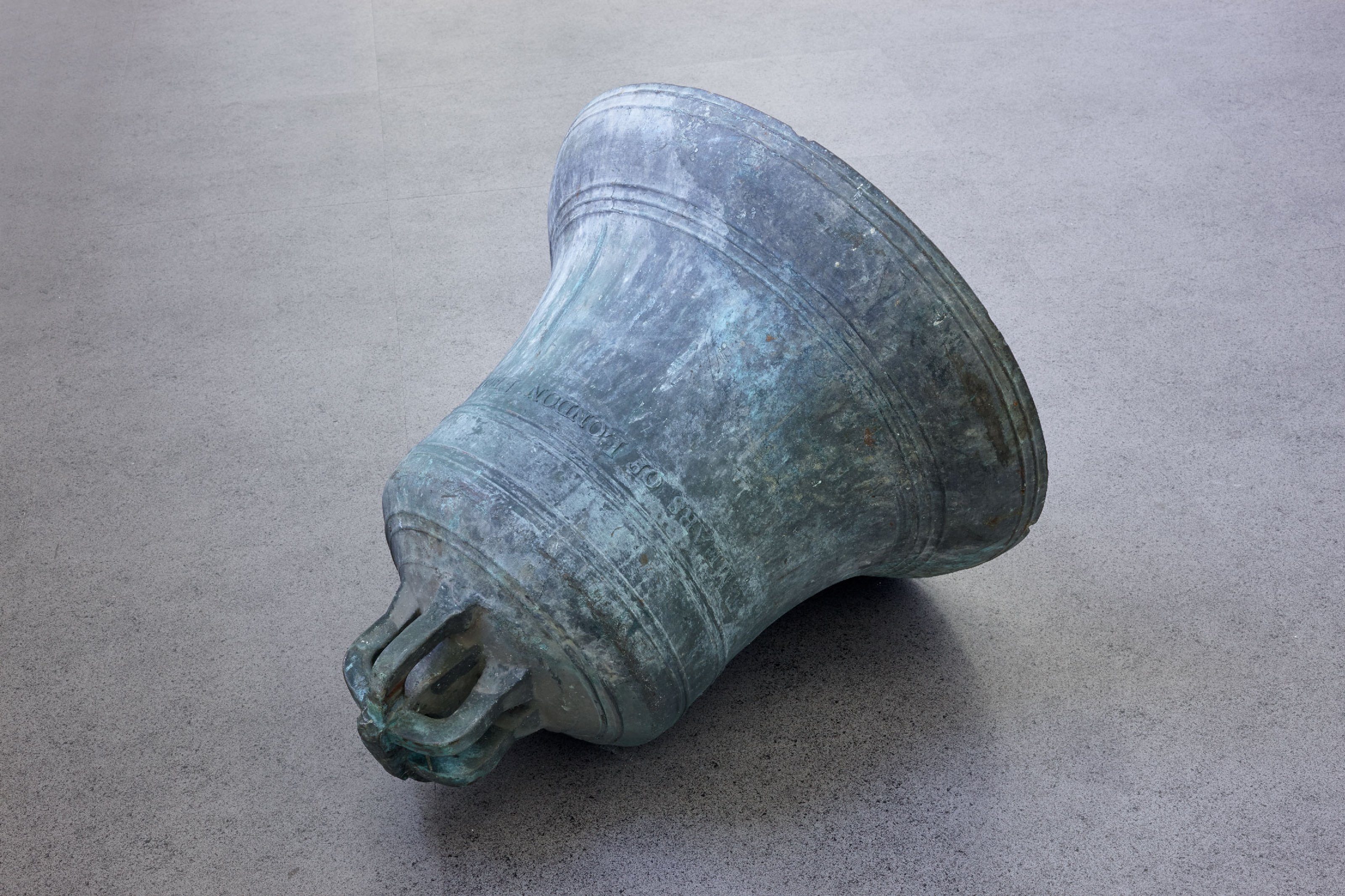 A big, metal bell on a gray long corridor of an art gallery.