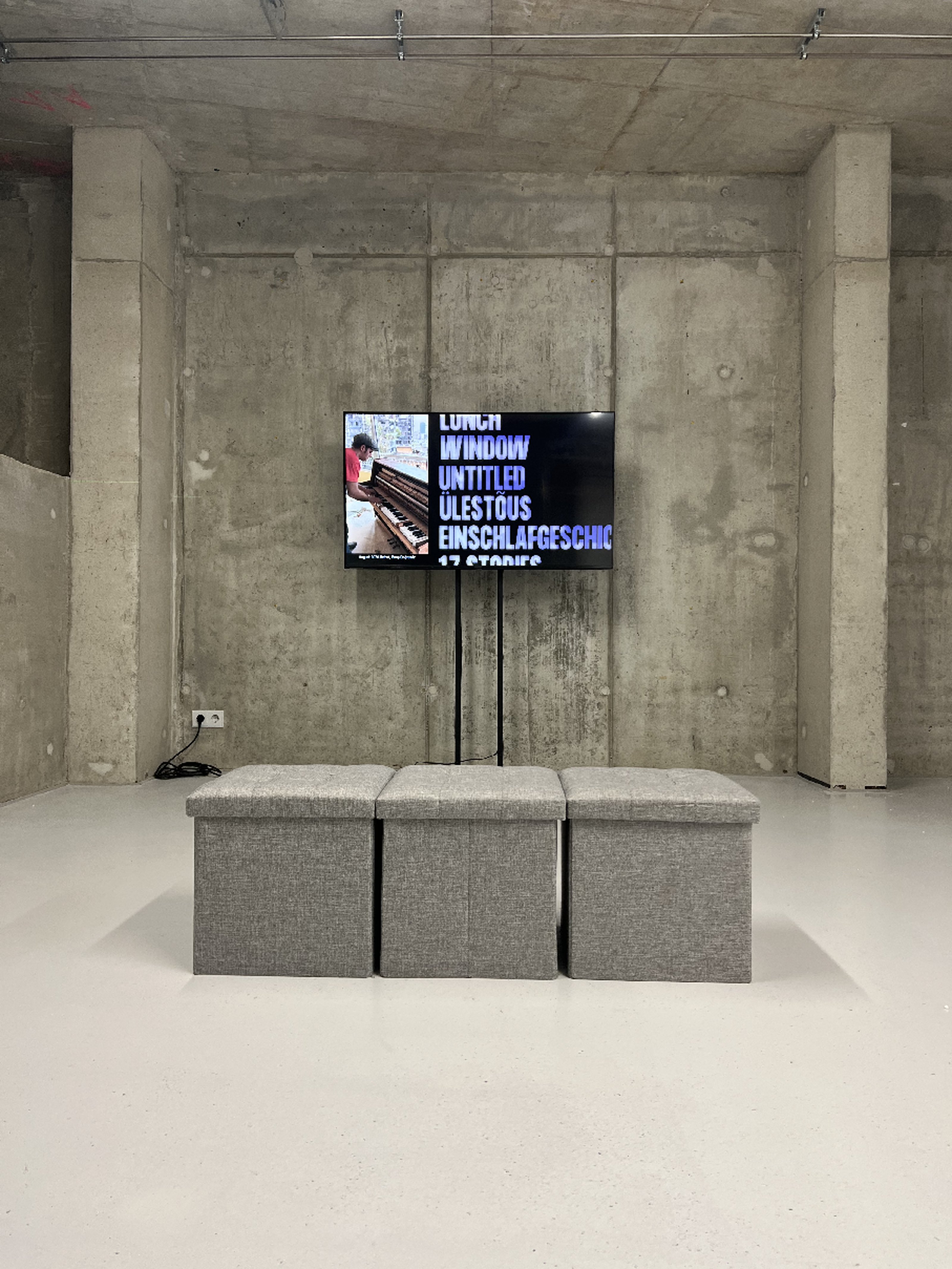Three stools in front of a video placed on the wall of a concrete-walled gallery.