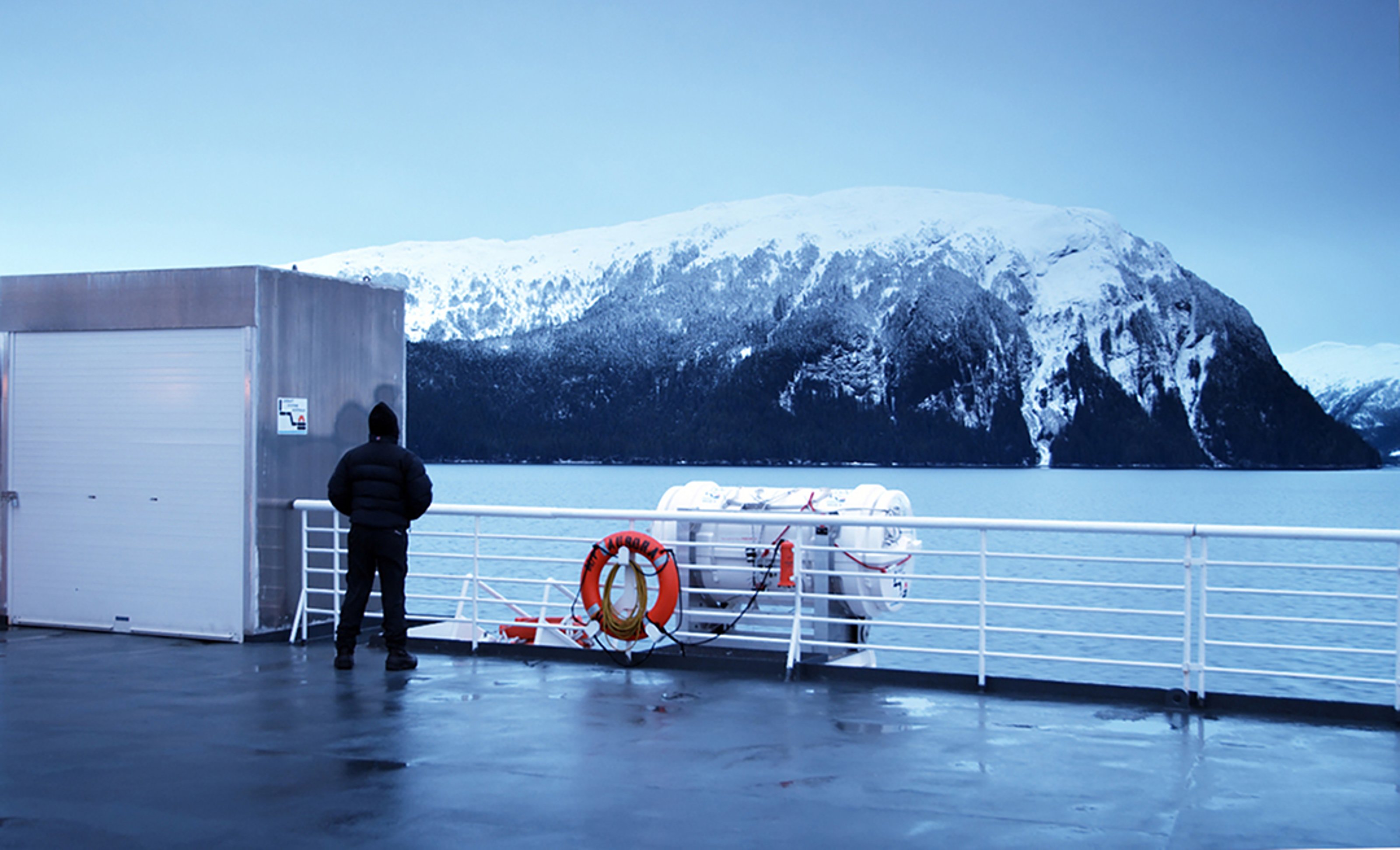 A man with his back turned to the camera, standing on board of a ship. He is gazing towards a snowy mountain.