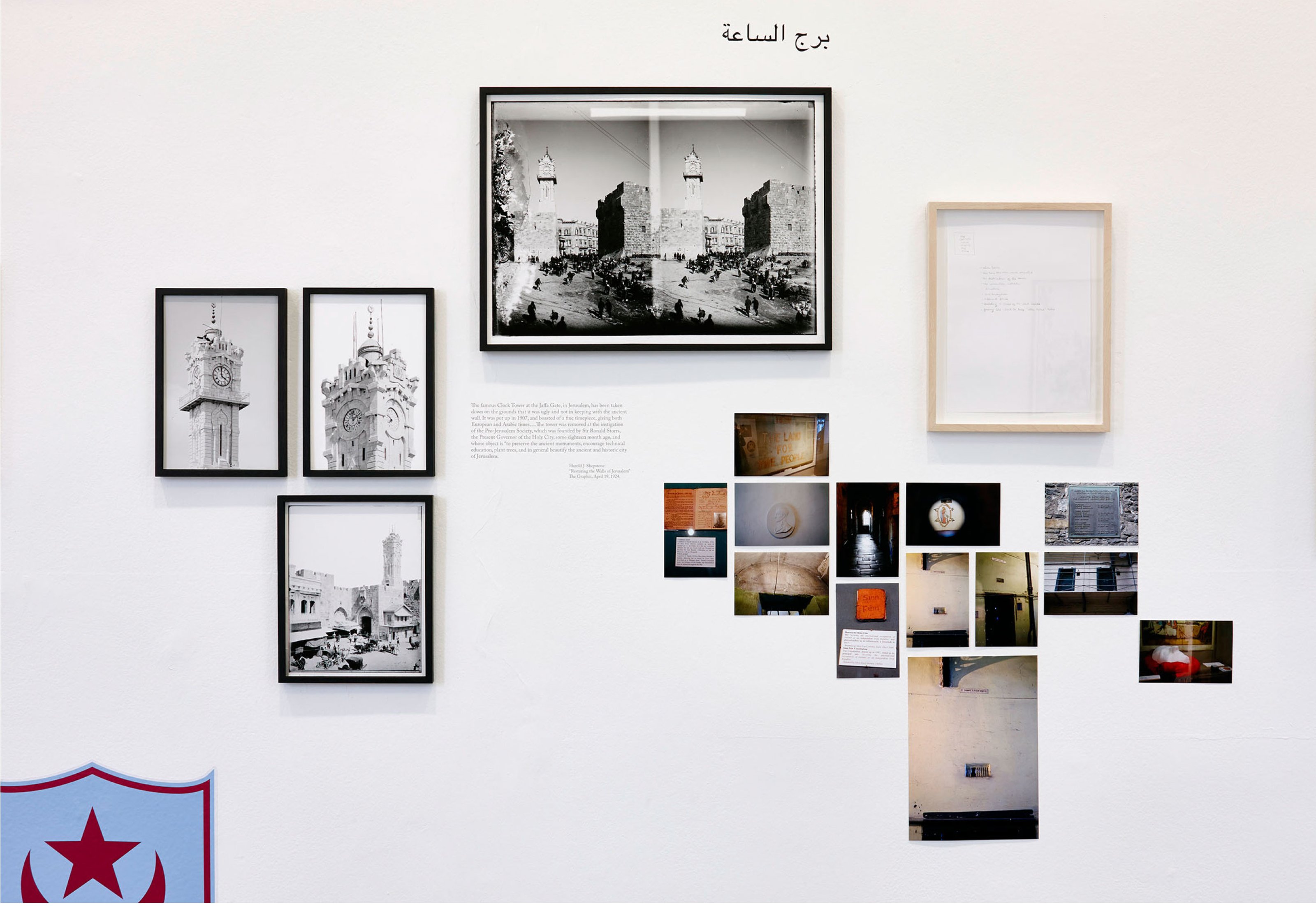 Various artworks displayed on a white gallery wall, including a black and white photo of the Jaffa clock tower among others.