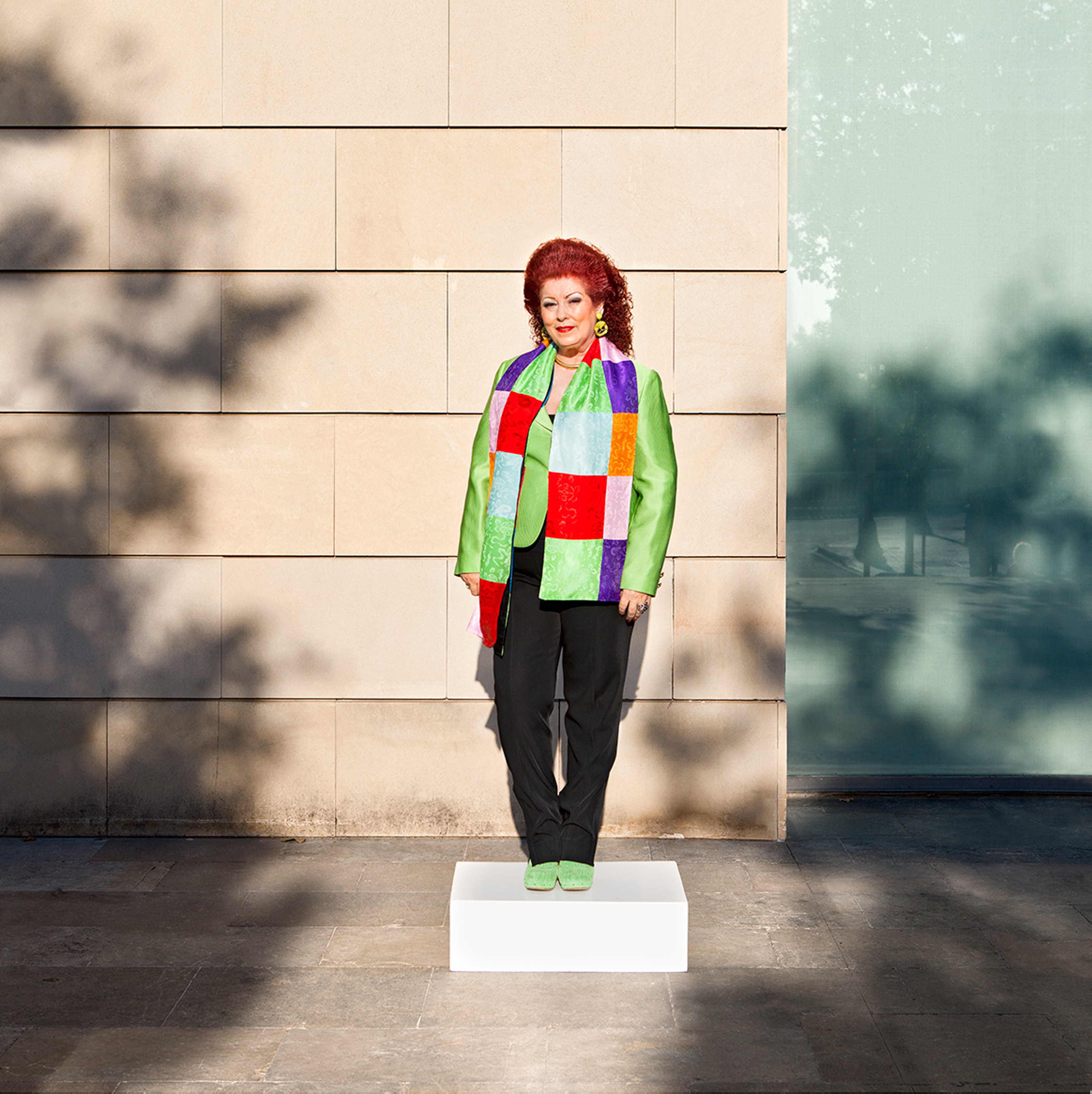 A middle-aged Consuelo Císcar with red wavy hair, green jacket and shoes, and black pants stands on a pedestal.