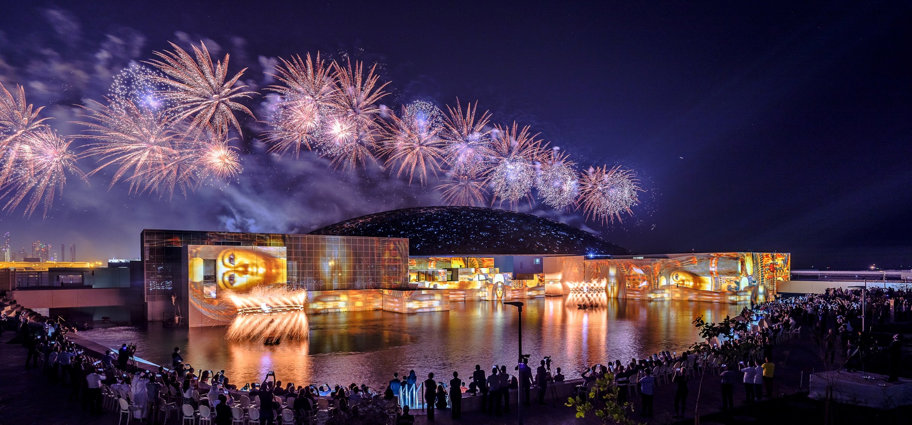 A huge crowd stands in front of the building at night during the celebration of the Louvre Abu Dhabi's opening with fireworks.