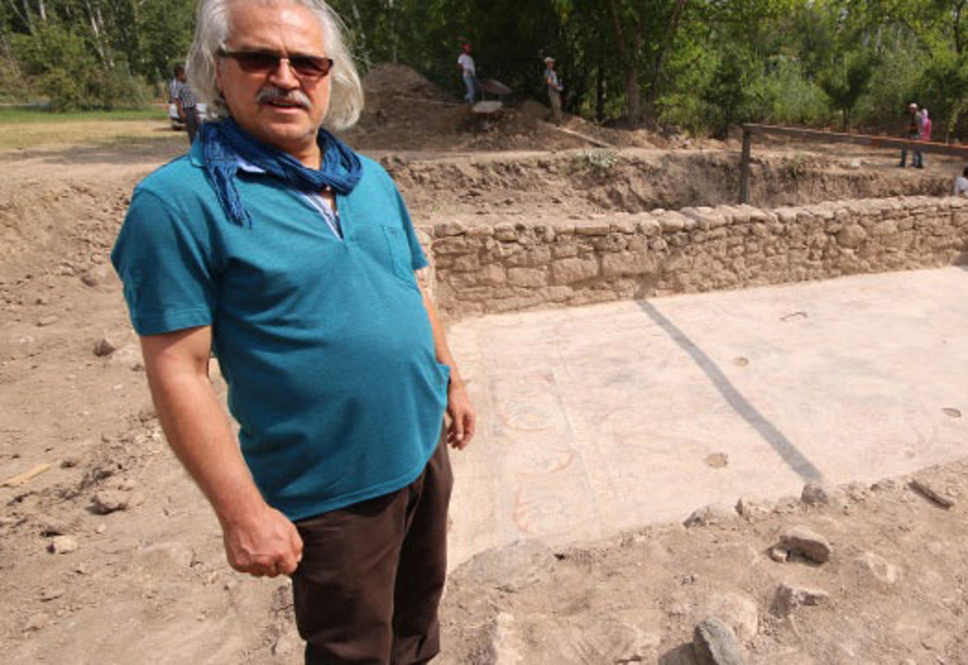 A white long-haired man in a blue shirt stands next to an archaeological dig.