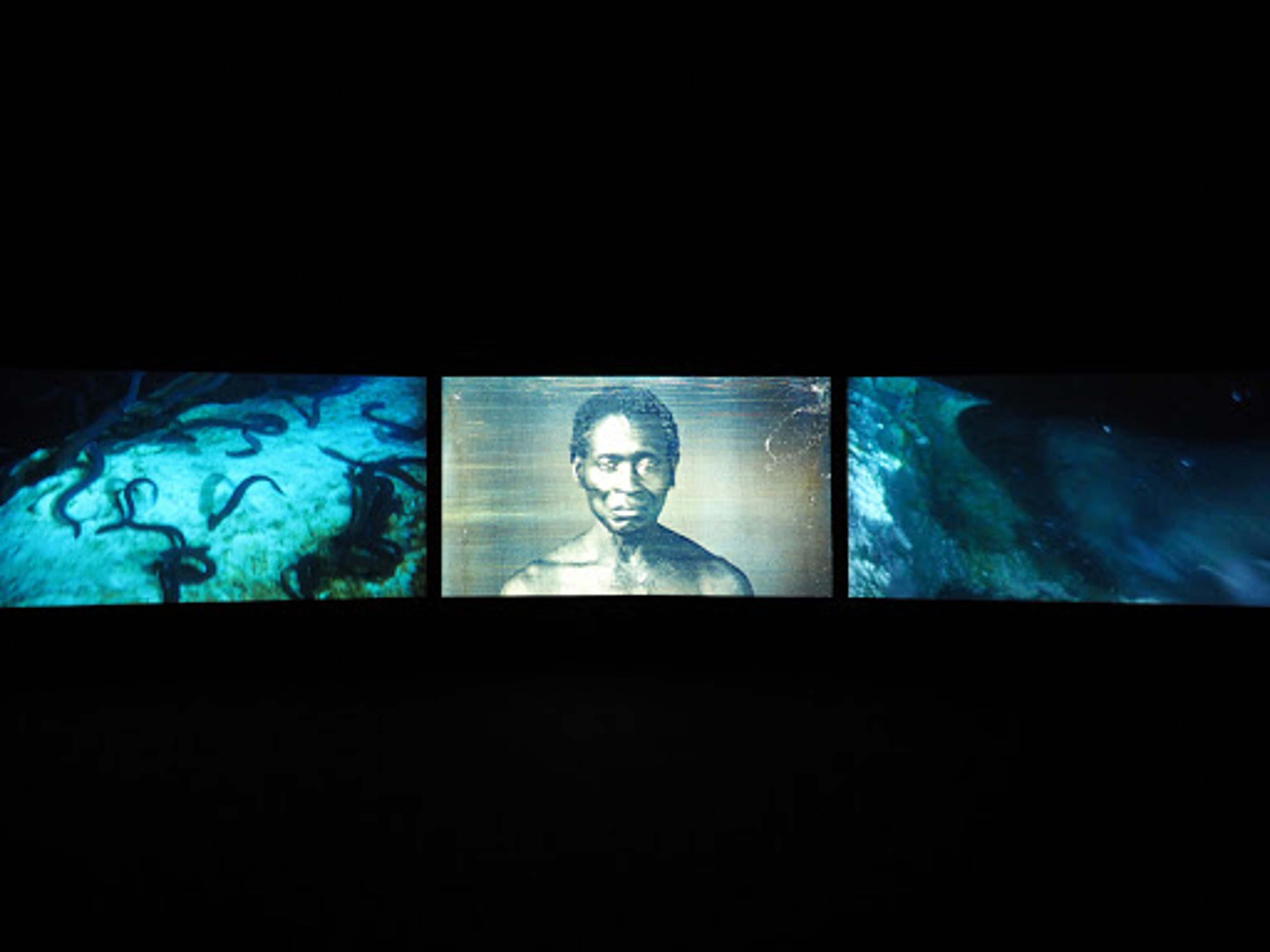 Video on three screens horizontally arranged. On the left and right screen are images of the ocean floor. At the center, an old photograph of an African person