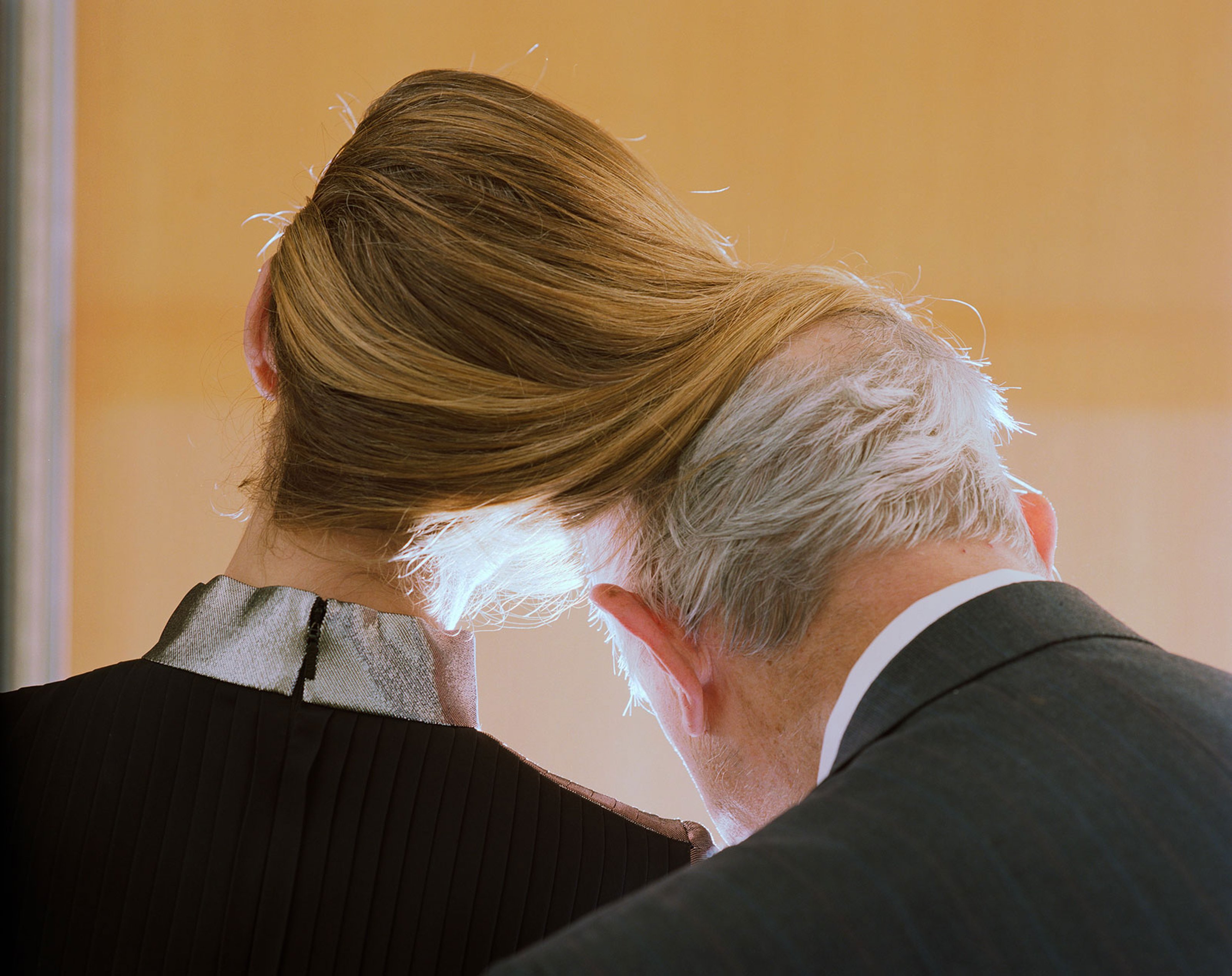 Looking at a woman and a man from the back. The woman's hair is wrapping around the man's, the man is older and slightly bald. Their heads are leaning towards each other