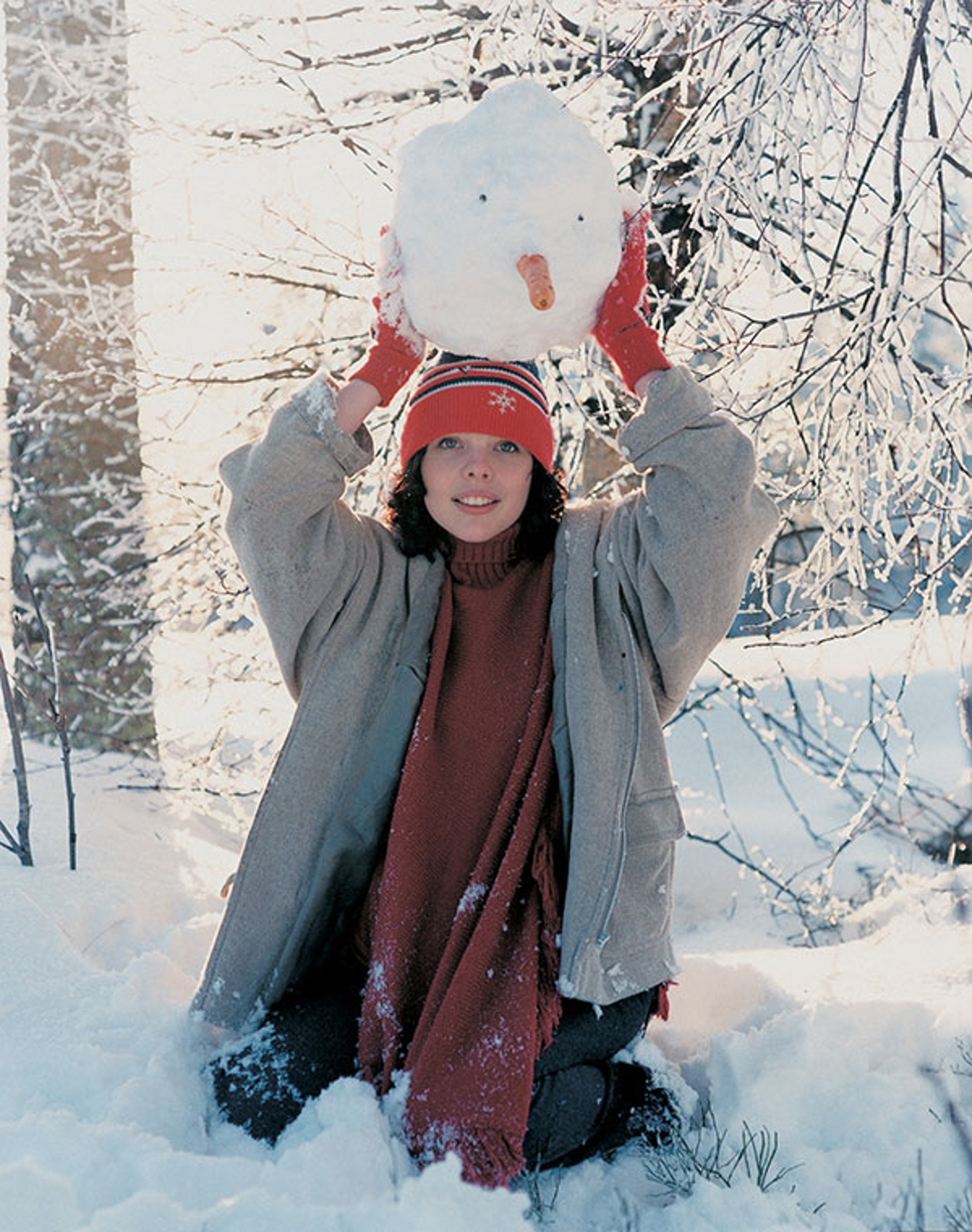 A woman sitting in snow, holding a snowman's head on top of hers.