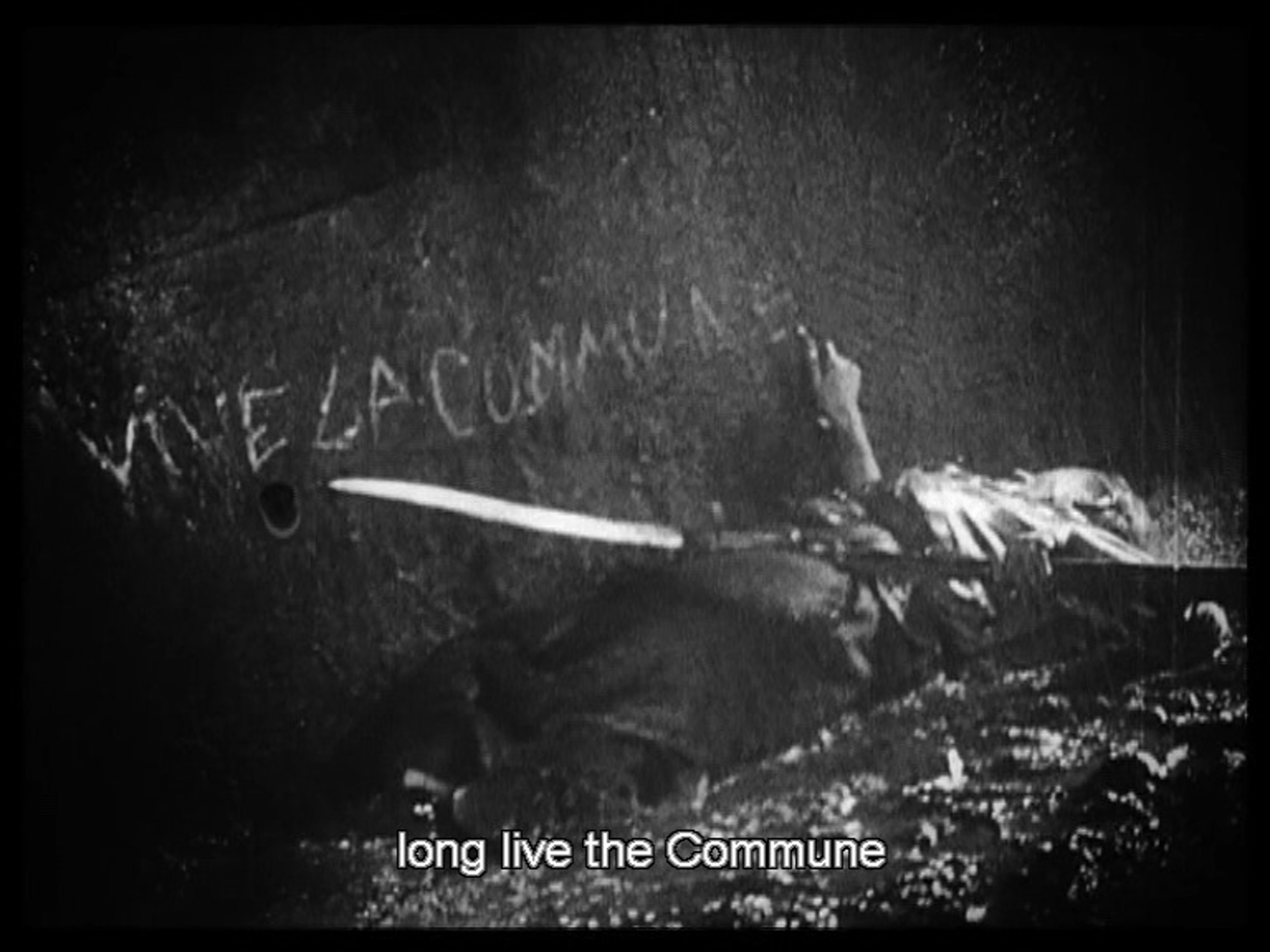Movie still, someone lying in the rain, possibly dying, while writing Vive La Commune (Long Live the Commune) on a wall