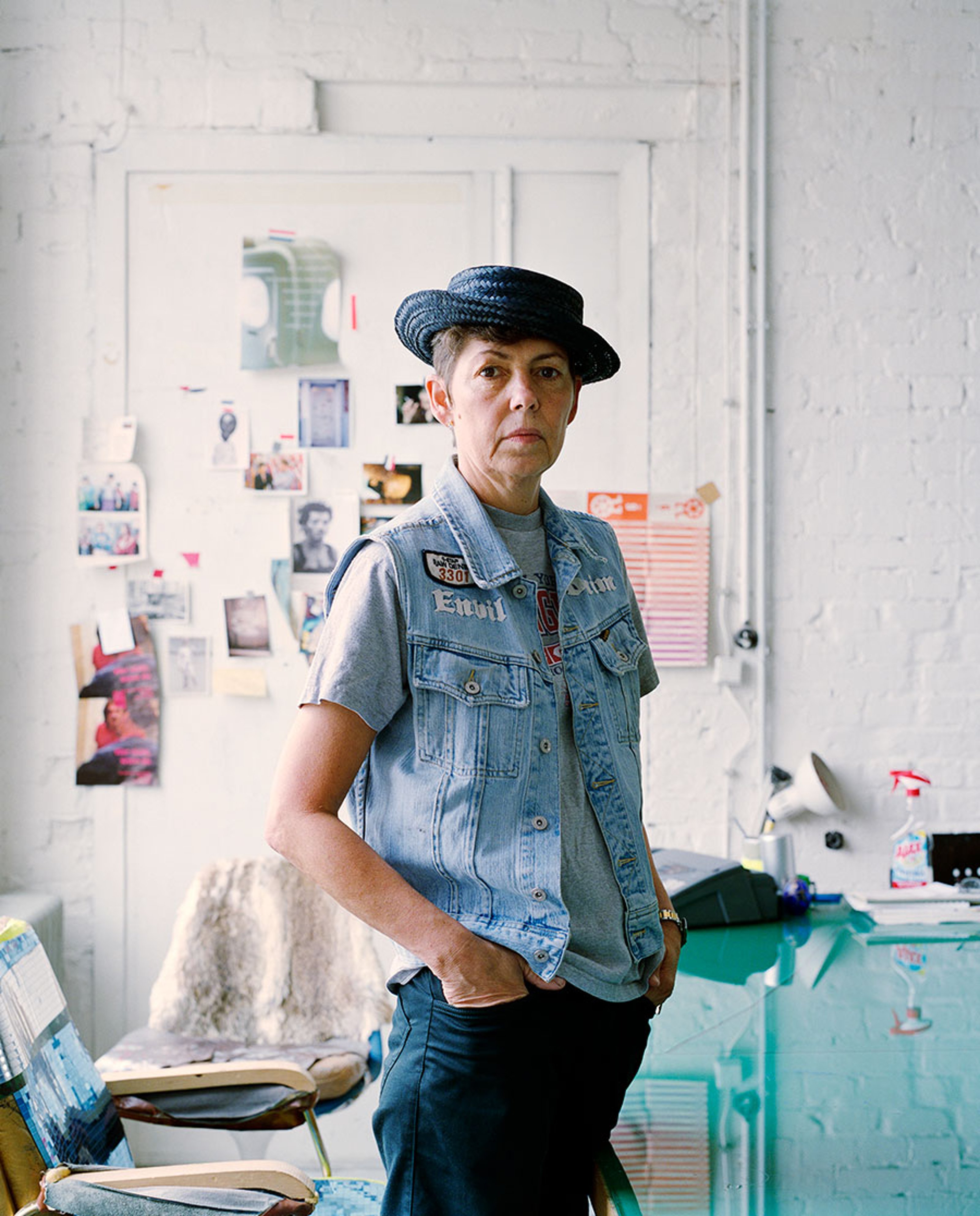 Photograph of Isa Genzken in her studio looking at the camera with a hat on and her hands in her pockets