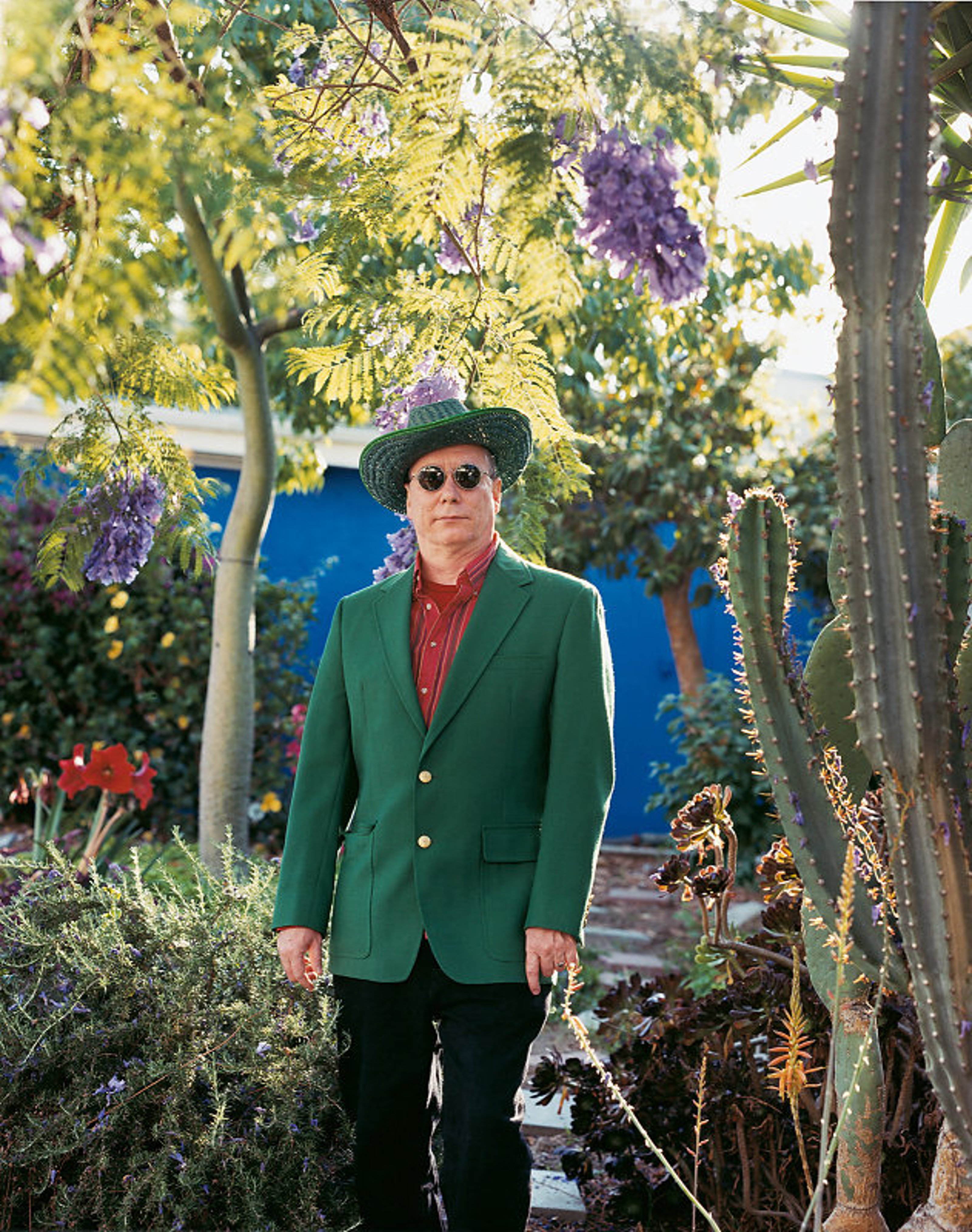 Mike Kelley in a colorful suit and a hat, standing among trees and cacti in Los Angeles