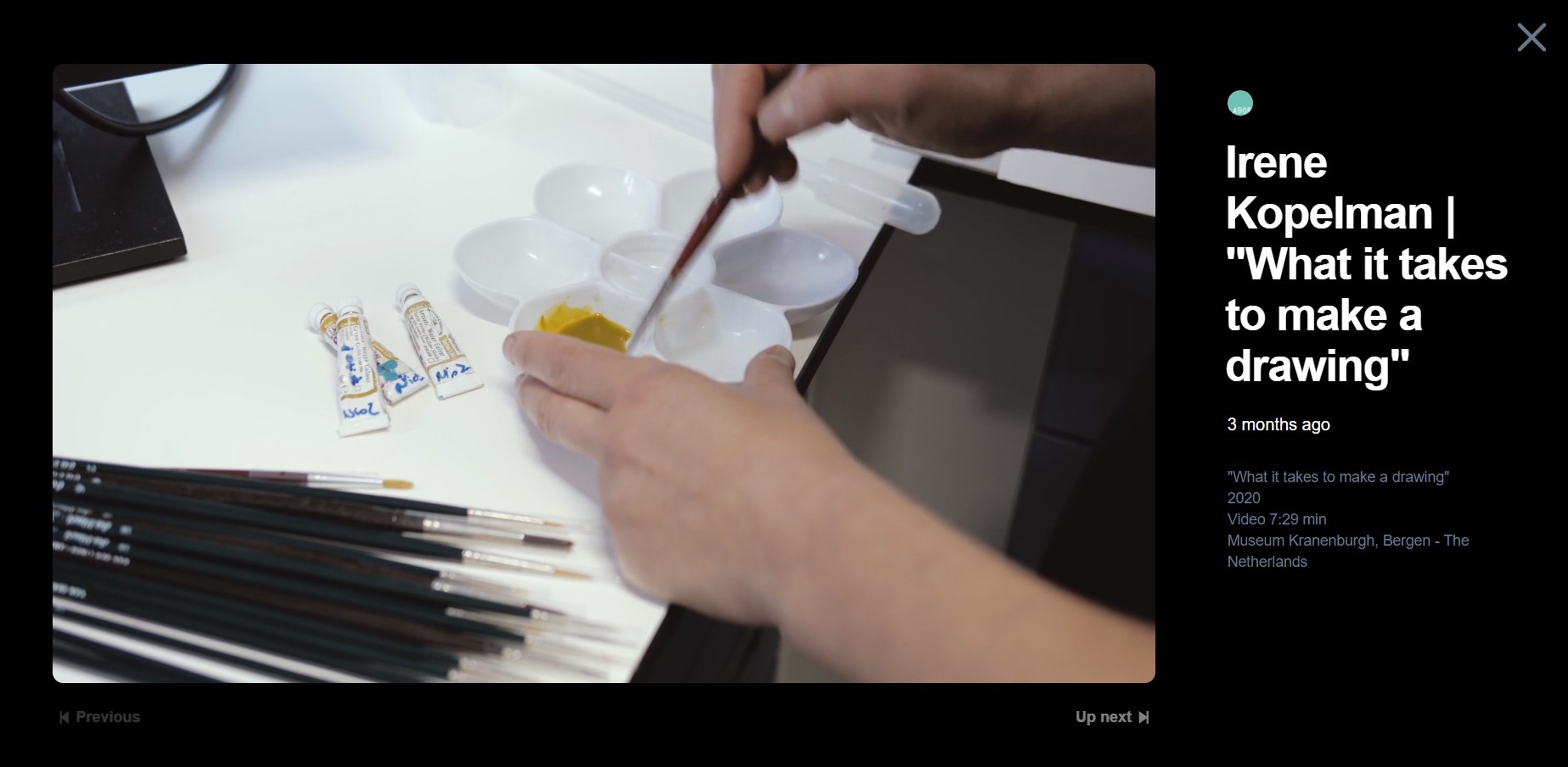 Still image from a video. A hand holding a brush, mixing yellow paint in a palette.