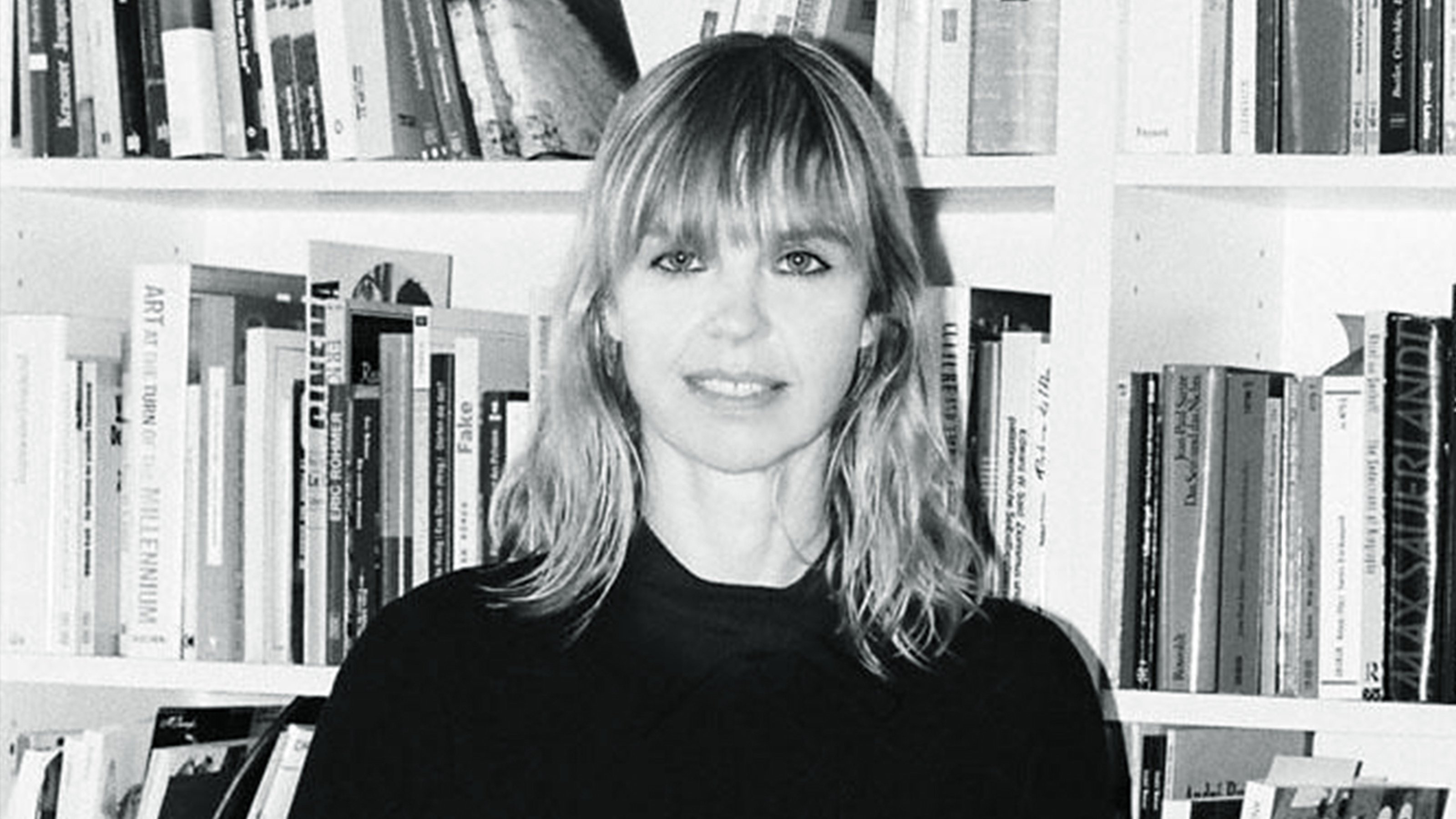 Black and white image of Isabella Graw standing in front of a bookcase and smiling into the camera