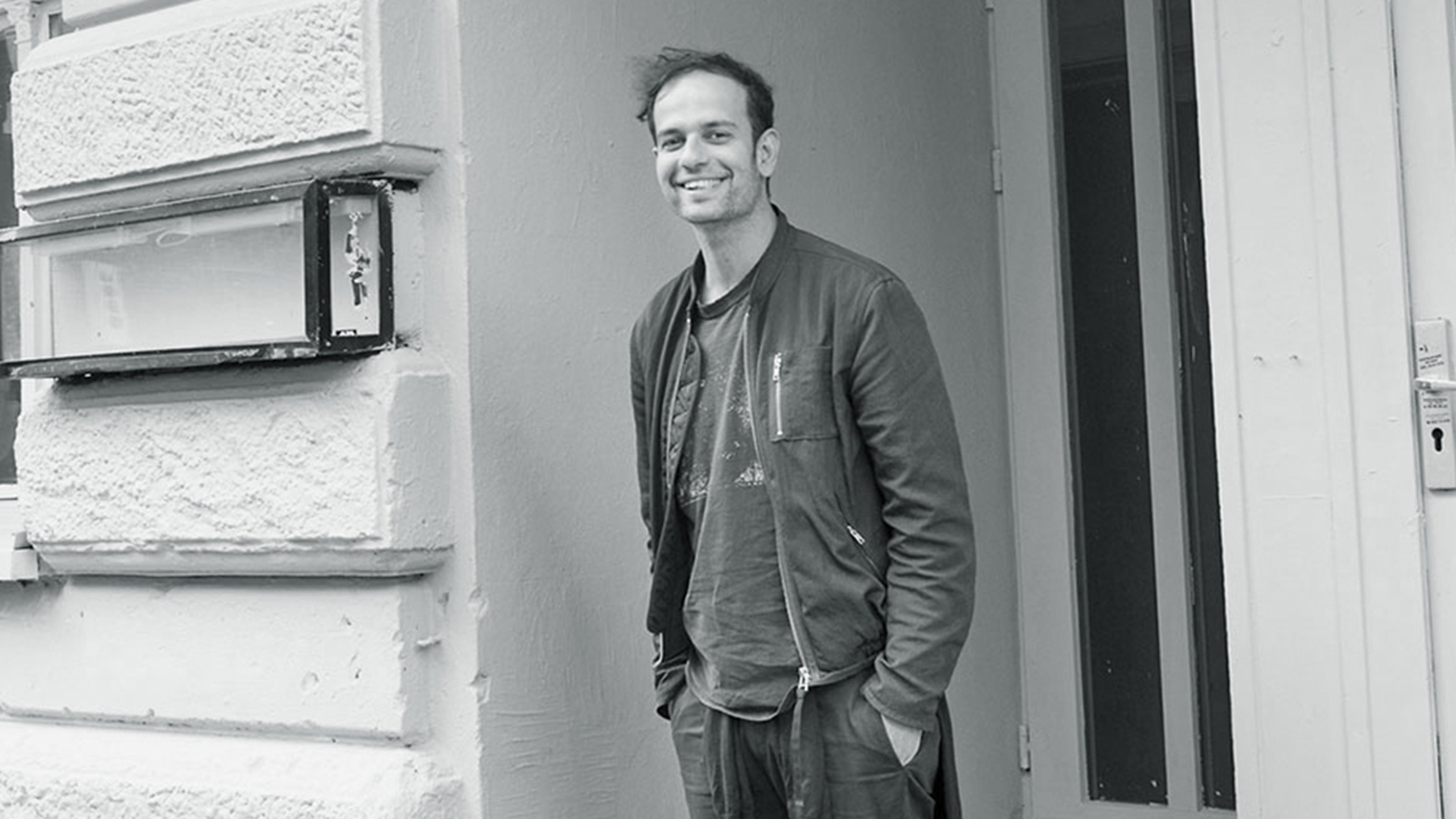 Black and white image of Tino Sehgal standing in front of a building and smiling at the camera