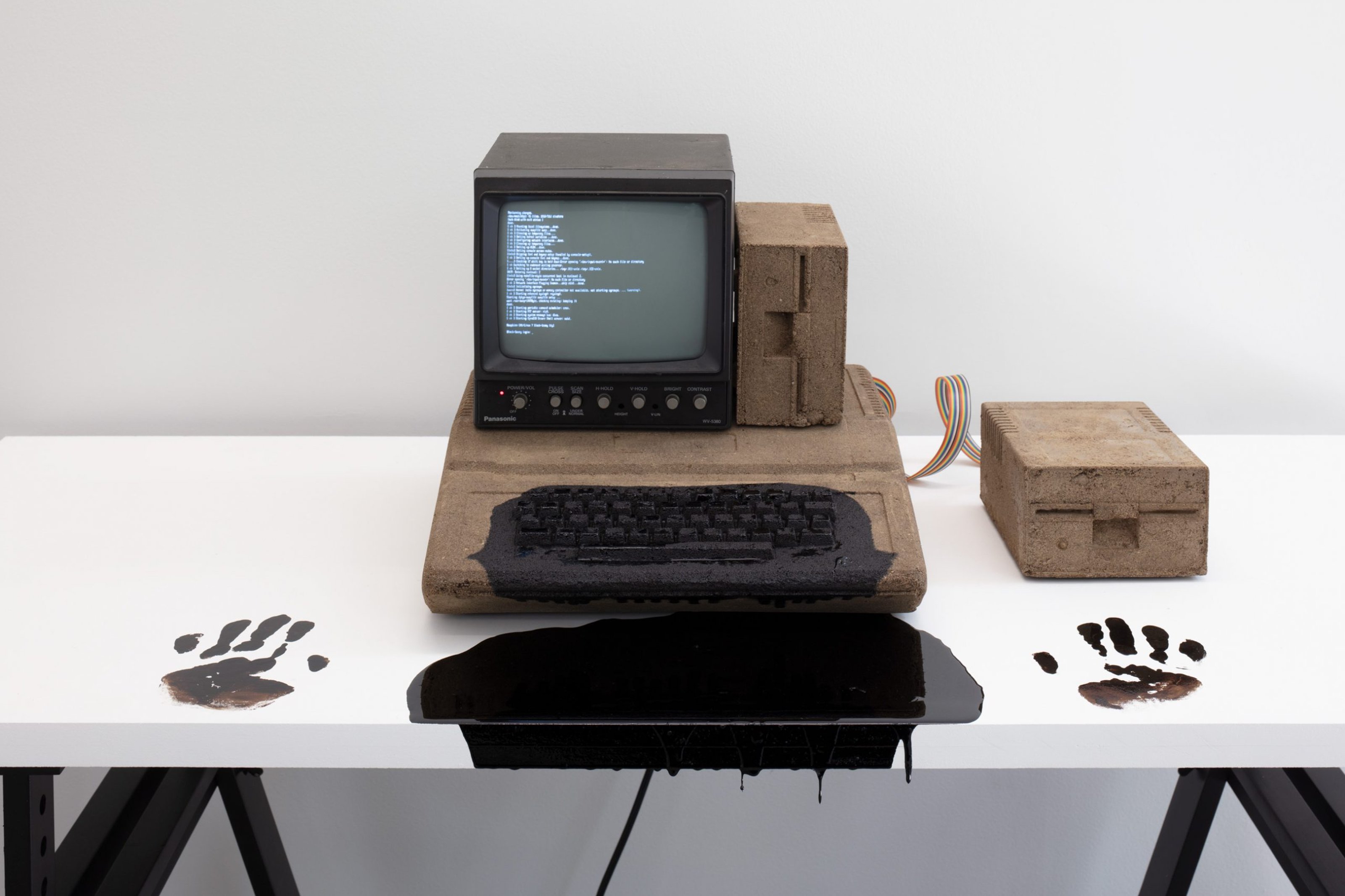 An old monochrome CRT monitor with keyboard, installed on a table. There is operating system running, and a black goo is coming out of the keyboards. There are hand prints on either side of the keyboard, marked with the black goo.